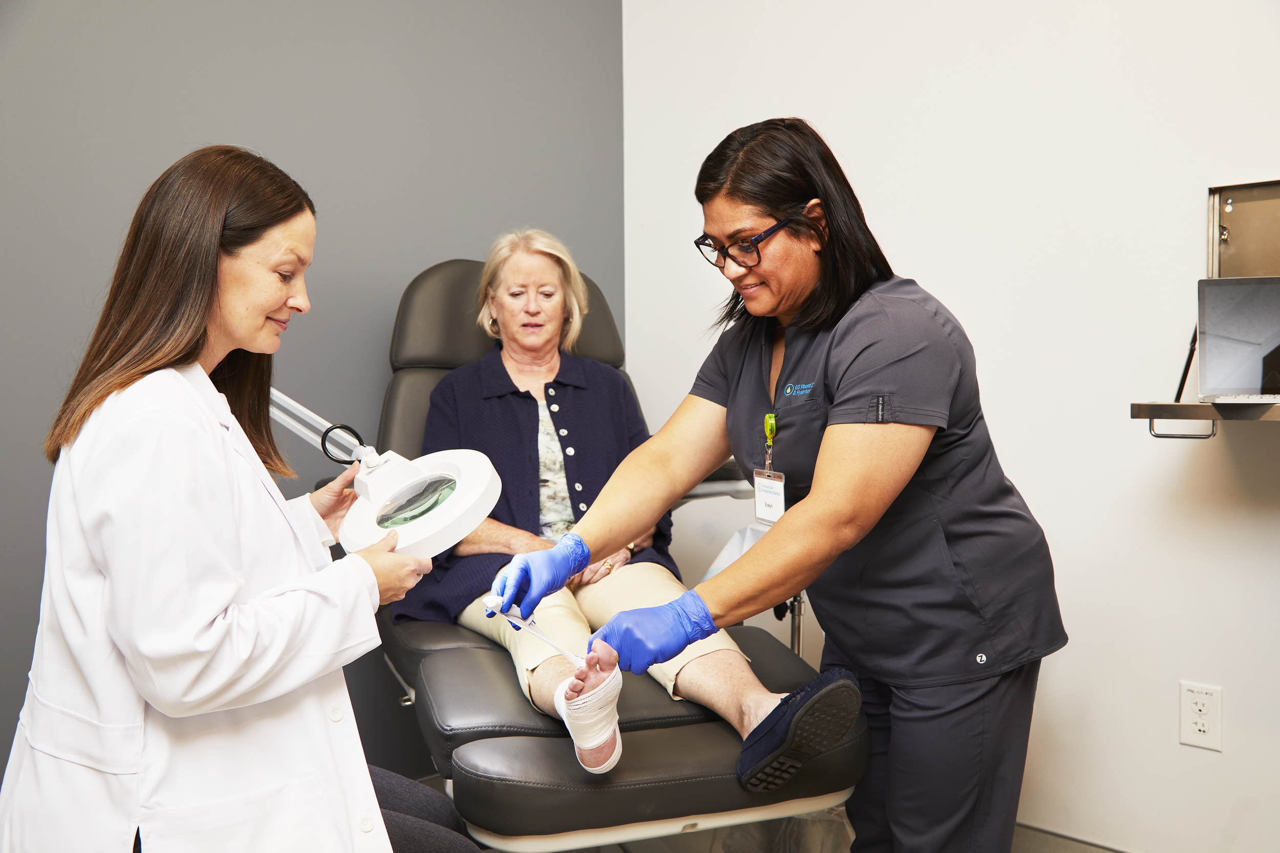 A medical professional giving an injection to a patient seated on a black medical chair, with another healthcare worker holding a magnifying lamp nearby, in a clinical setting.