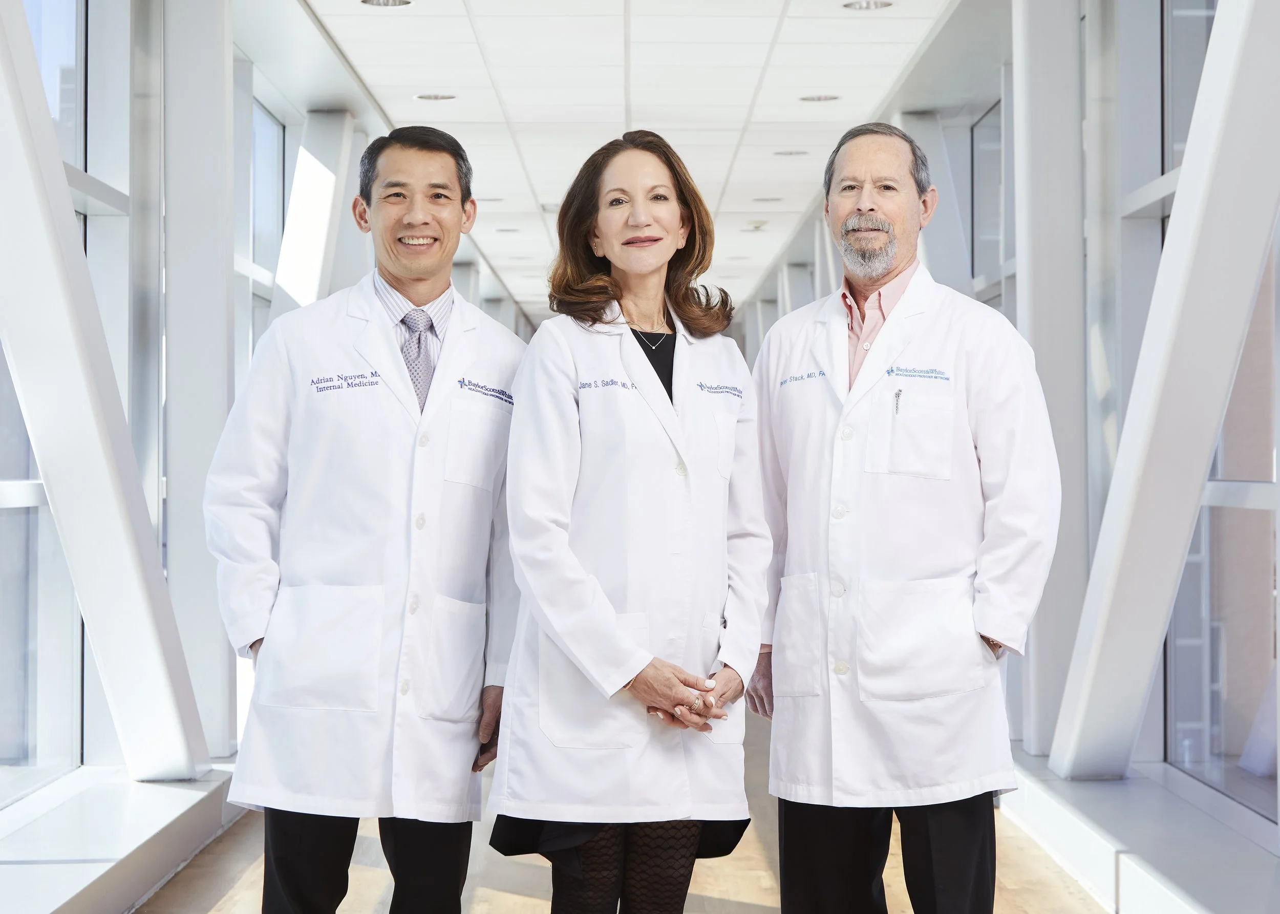 Three medical professionals wearing white coats standing in a modern hallway, smiling at the camera.