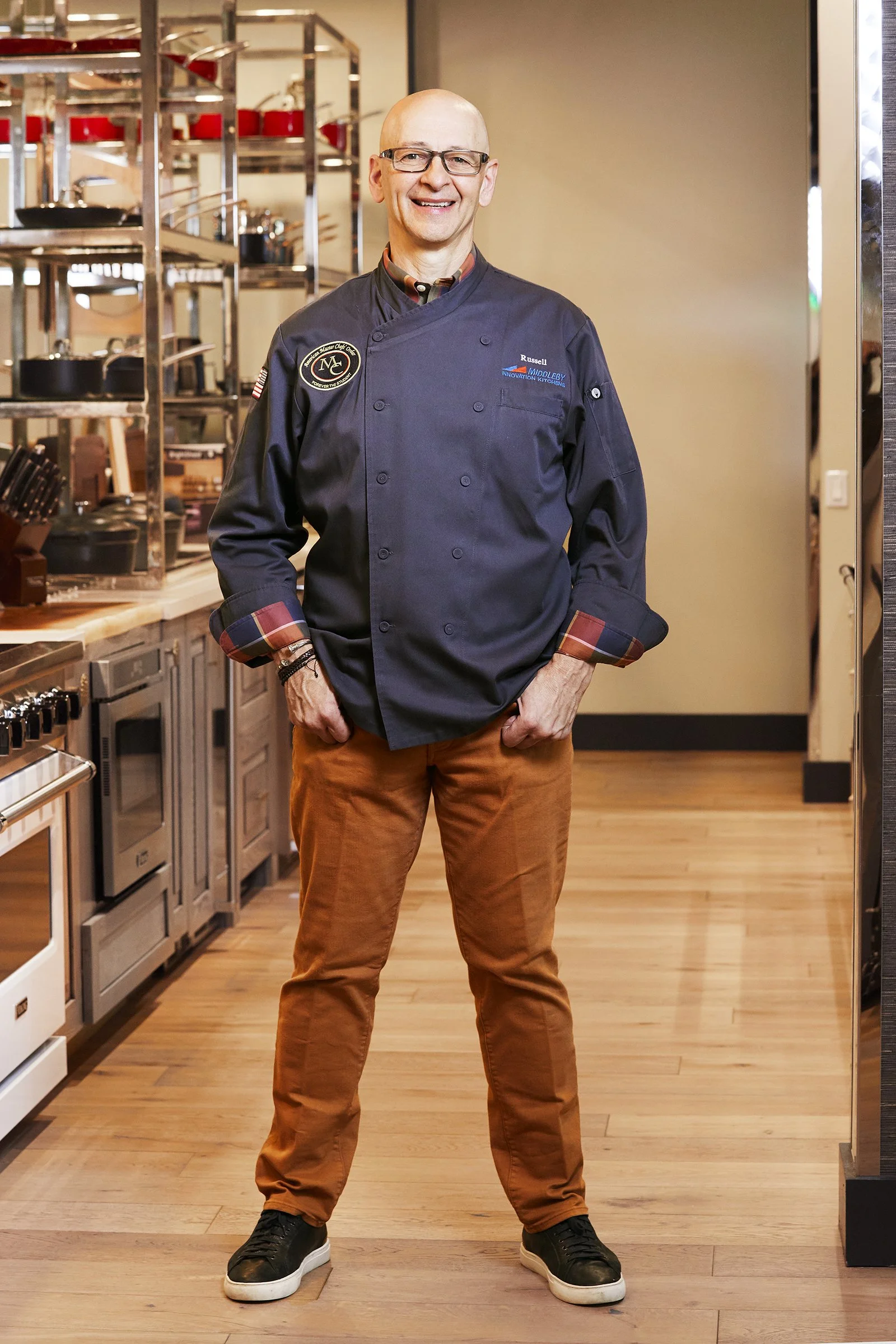 A man in a chef's uniform standing in a professional kitchen with cookware and appliances in the background.
