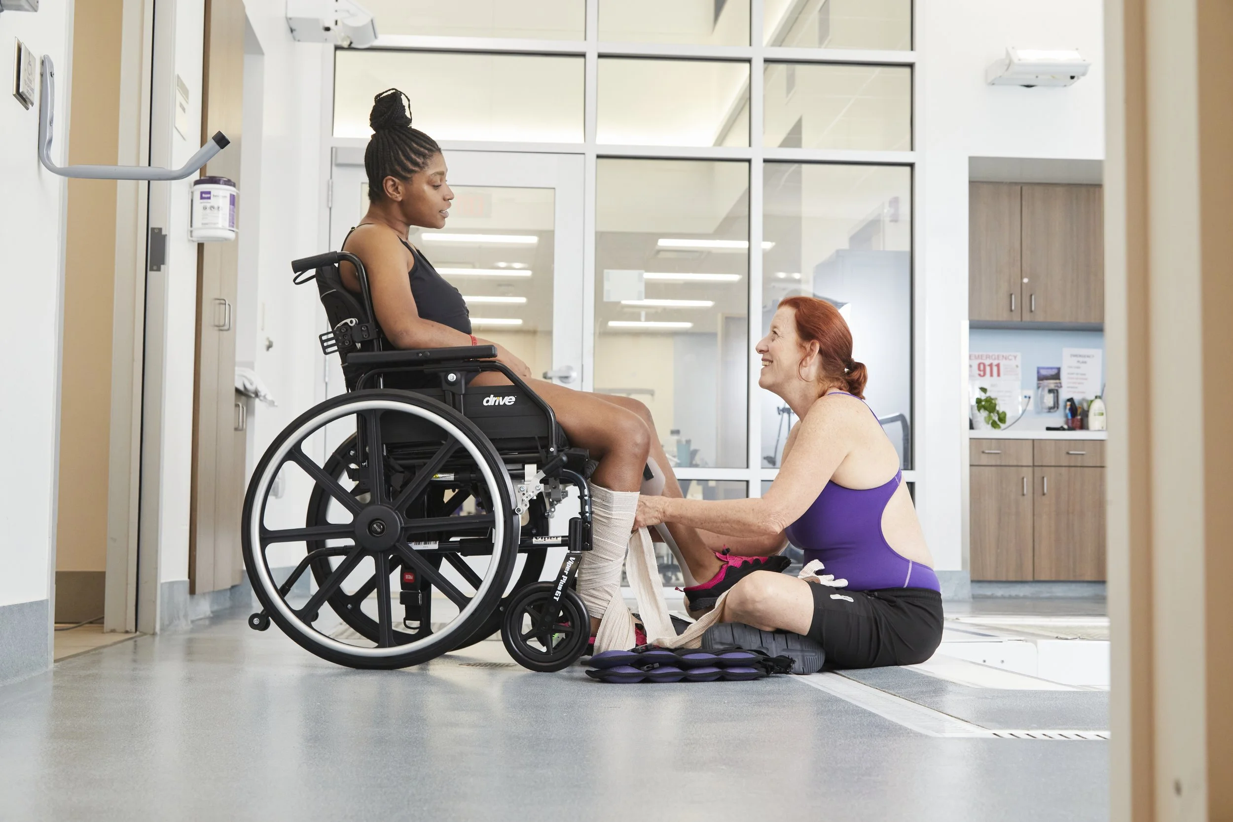 A woman in a wheelchair has her leg bandaged by a therapist kneeling on the floor in a medical facility.