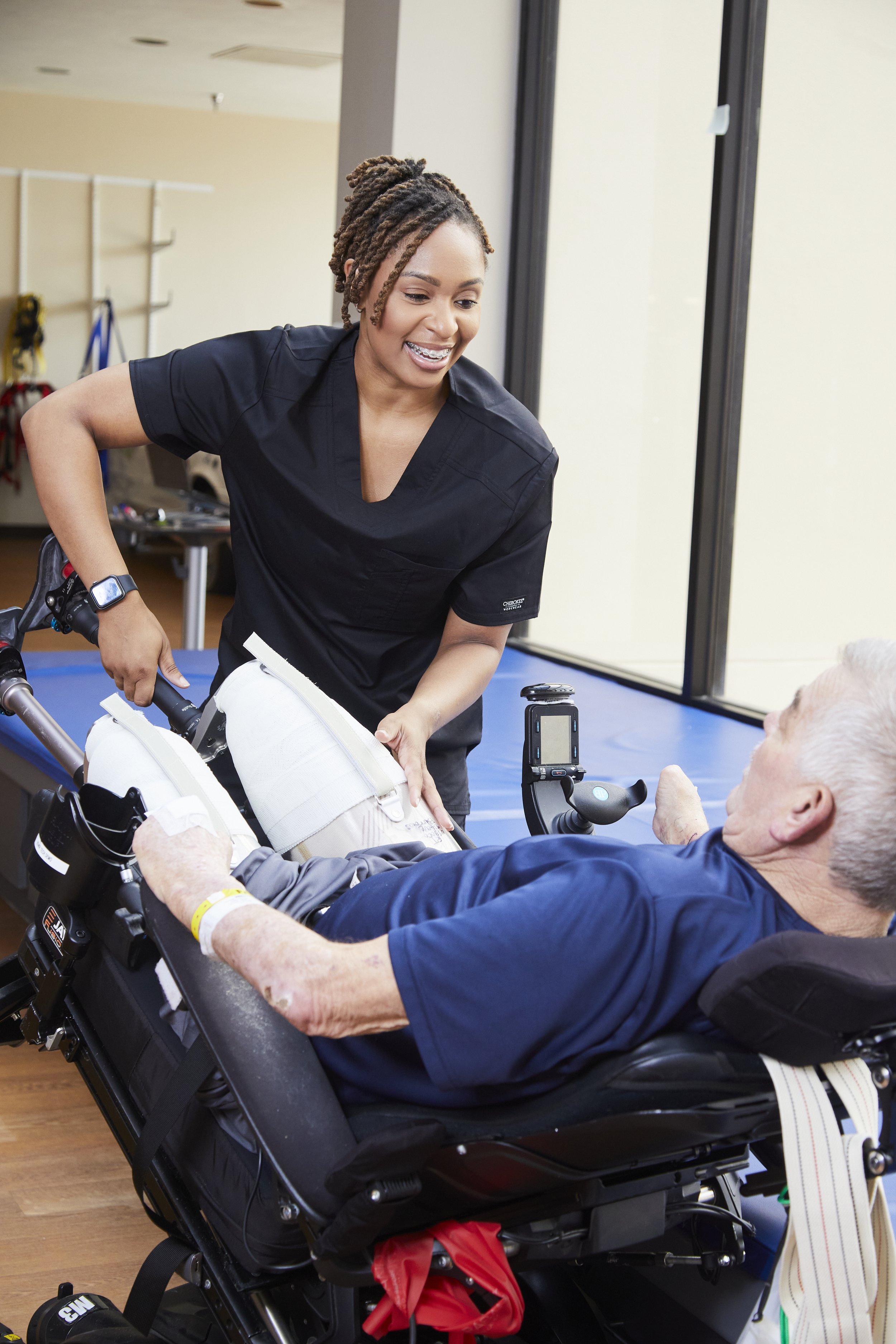 A female healthcare worker smiling at an elderly male patient in a wheelchair, adjusting his leg.