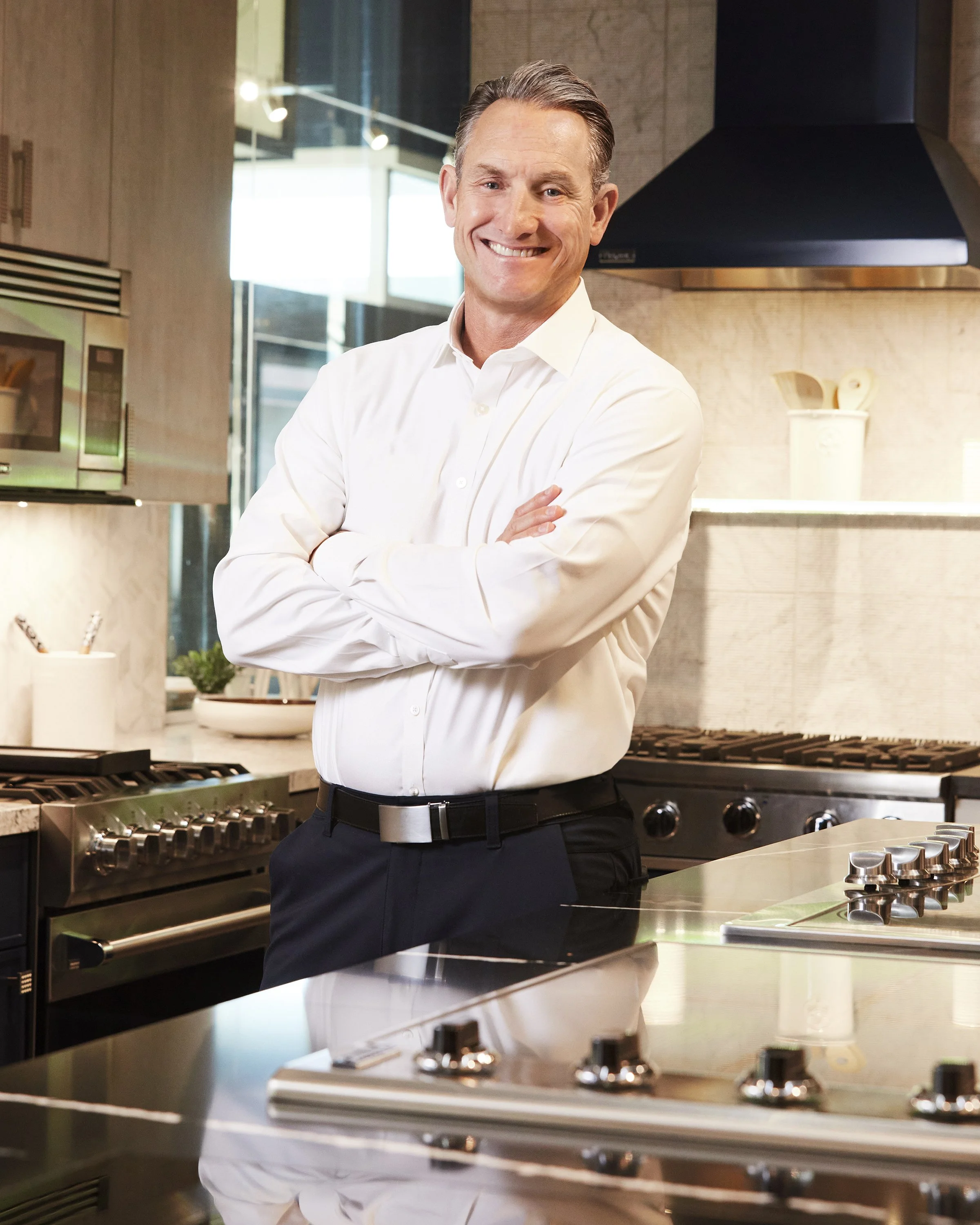 A smiling middle-aged man in a white dress shirt and black pants with a silver belt stands in a modern kitchen with crossed arms.