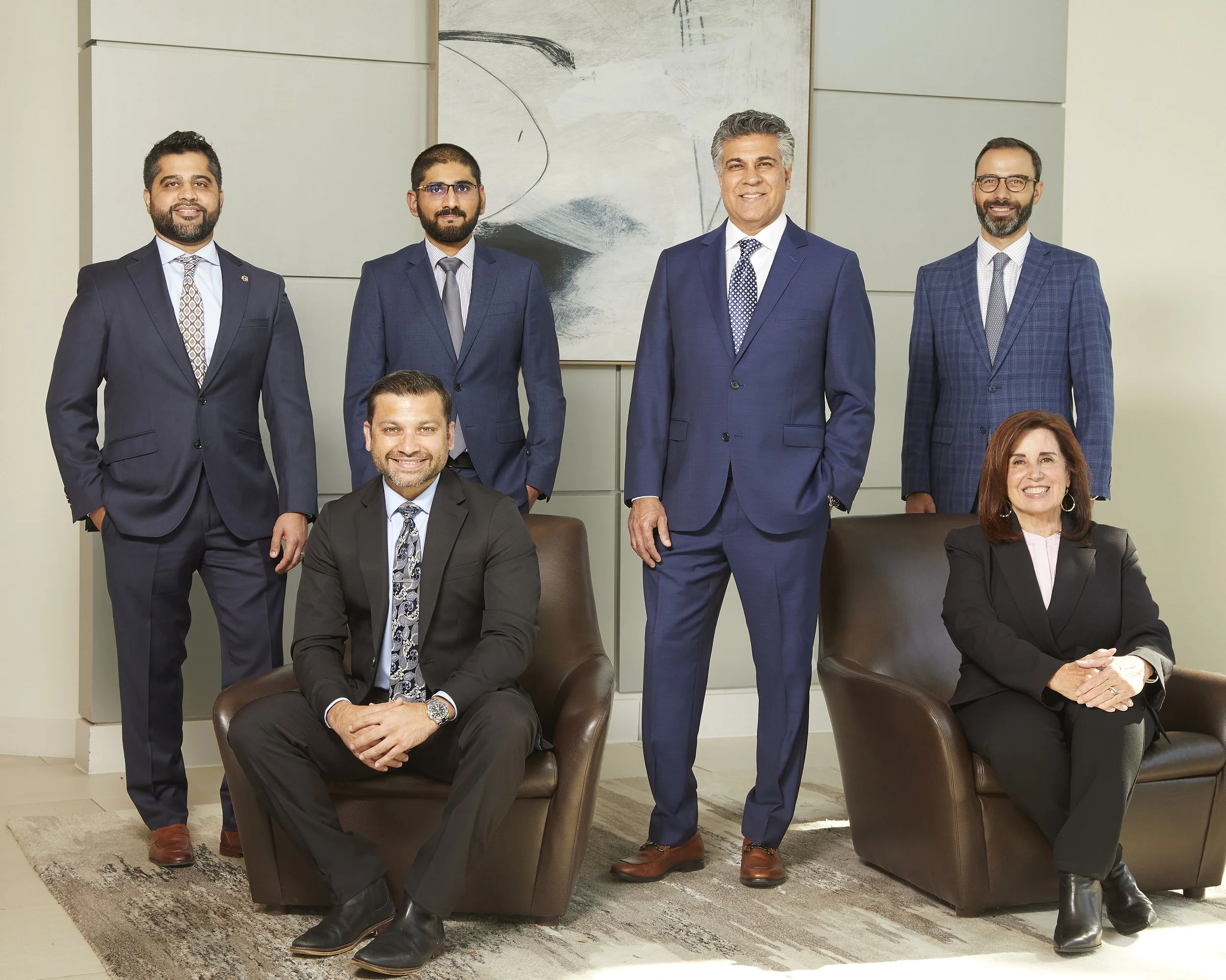 Group of seven professionally dressed business people in a modern office. Four men are standing behind two women sitting in leather chairs, and one man is seated in the middle of the front row.