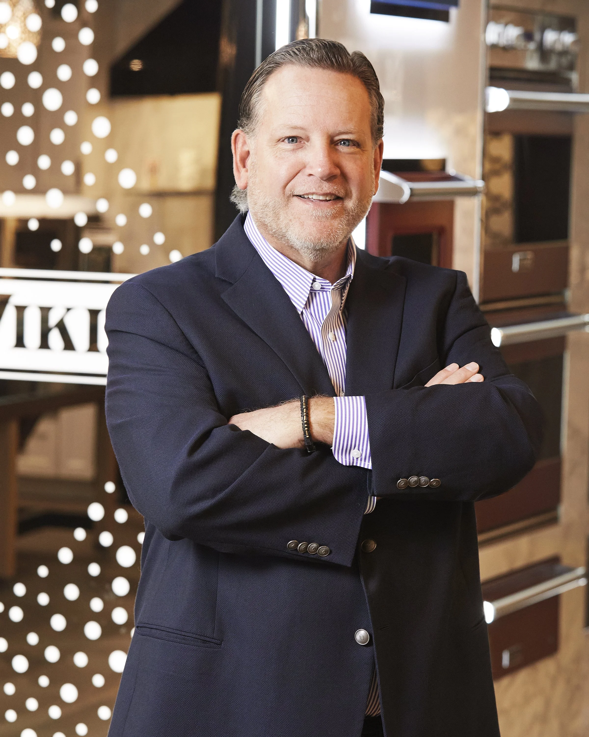 A middle-aged man with blond hair and a beard is smiling with arms crossed, wearing a dark suit jacket over a striped shirt, standing in a modern, stylish interior with shelves and decorative elements.