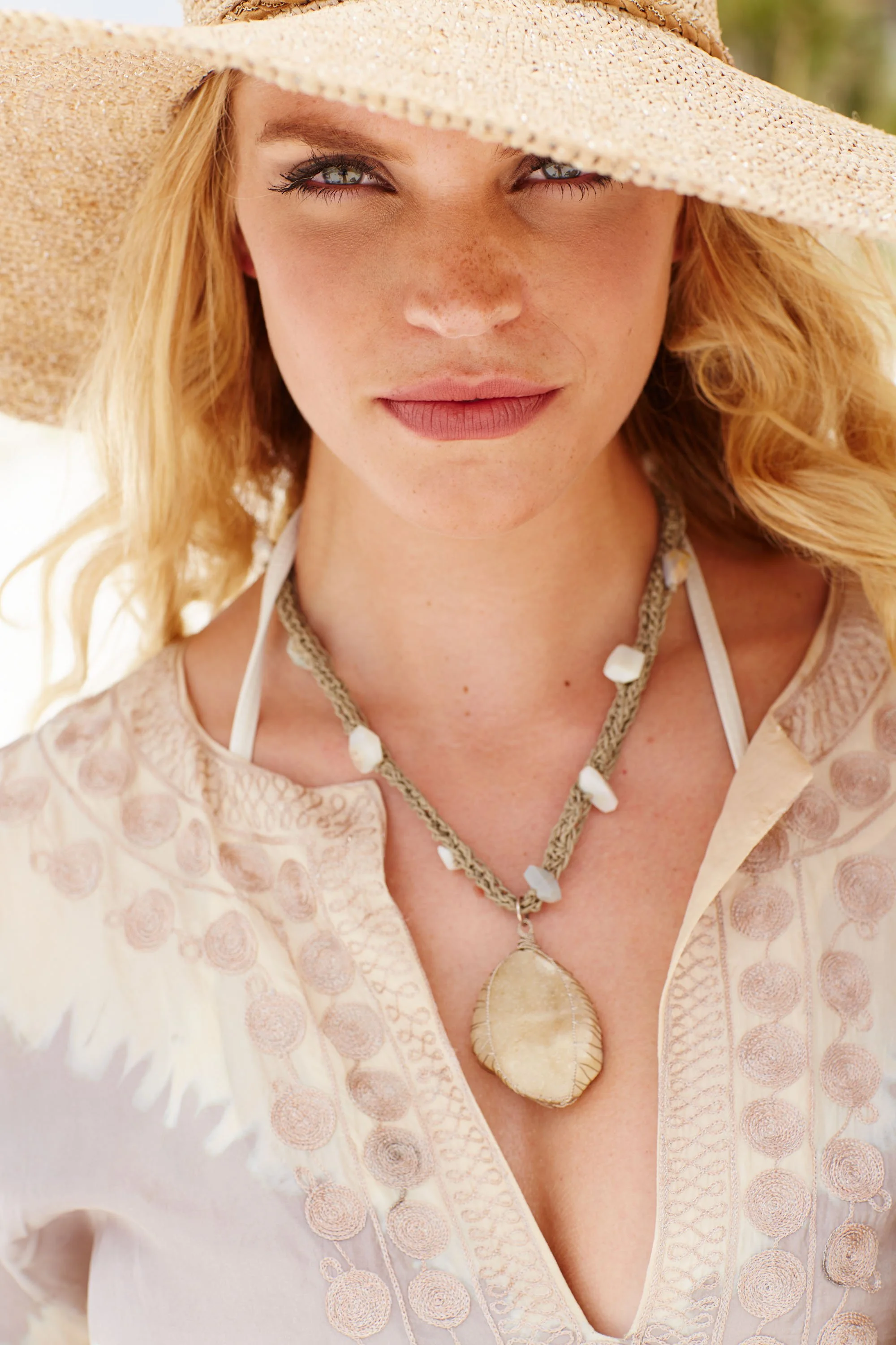A woman wearing a wide-brimmed straw hat, a patterned light-colored blouse, and a large shell necklace.