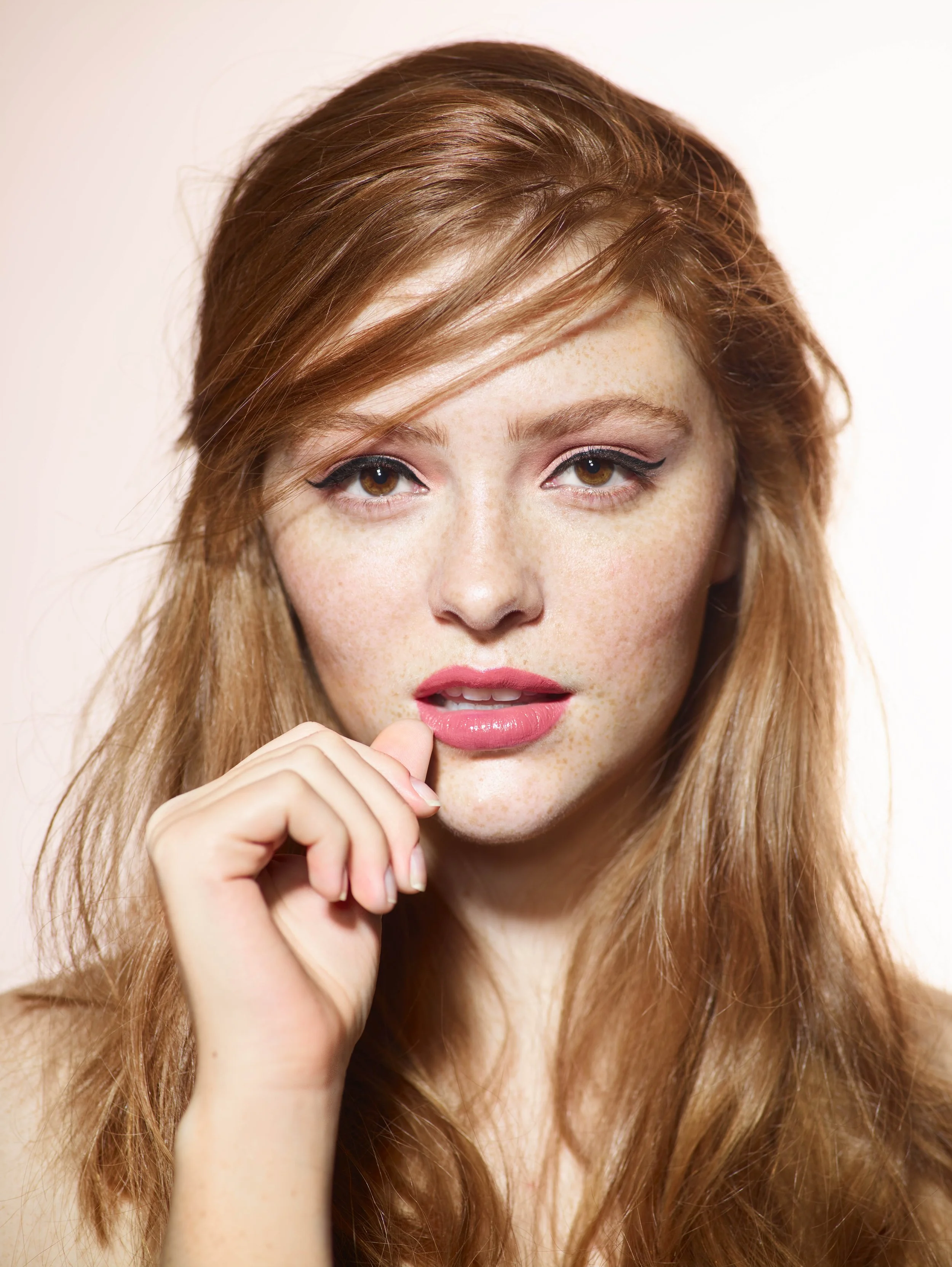 Close-up portrait of a young woman with red hair, light skin, and freckles, looking at the camera with a hand near her lips.