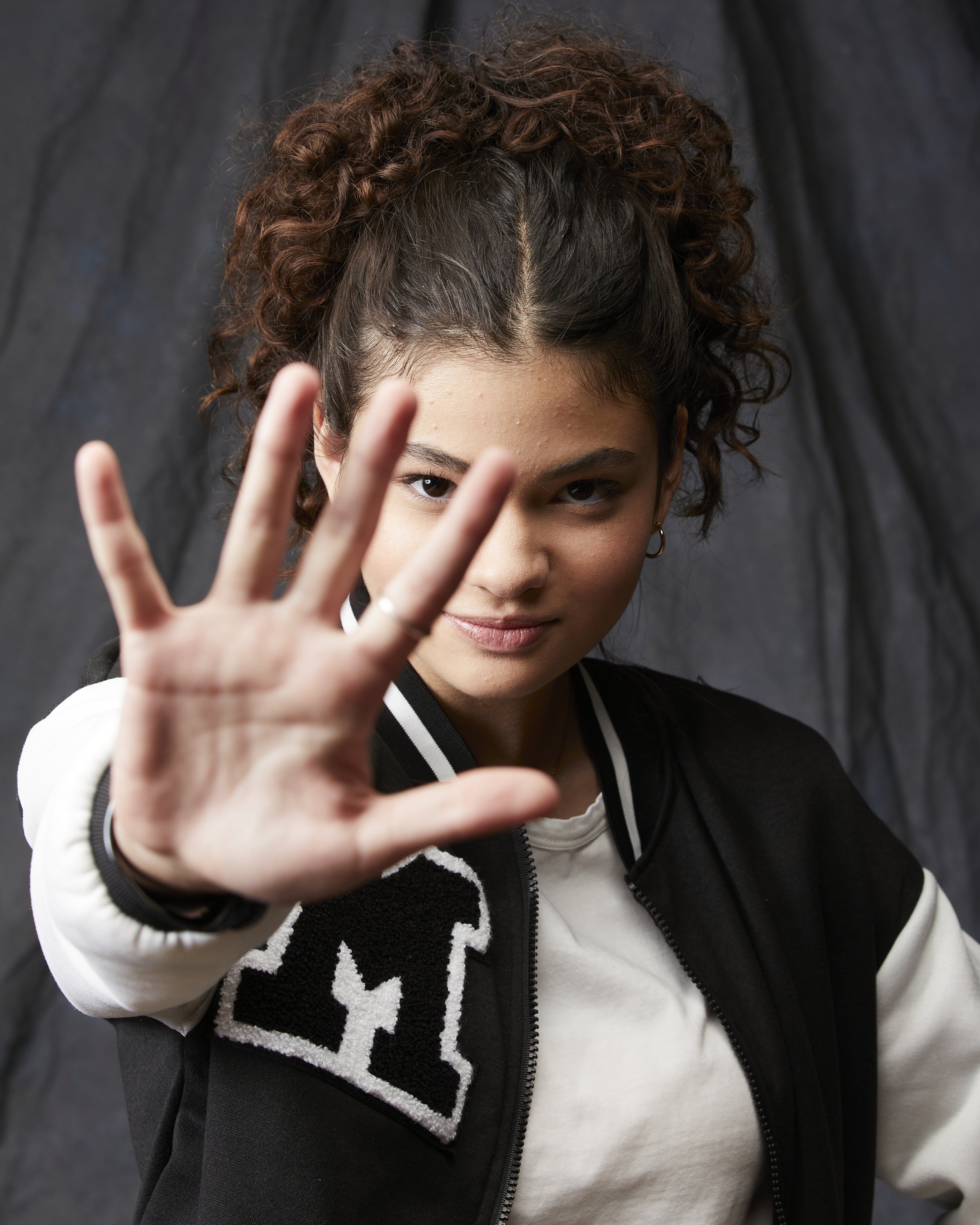 A young woman with curly hair extending her hand towards the camera, with a confident expression on her face, wearing a black and white jacket.