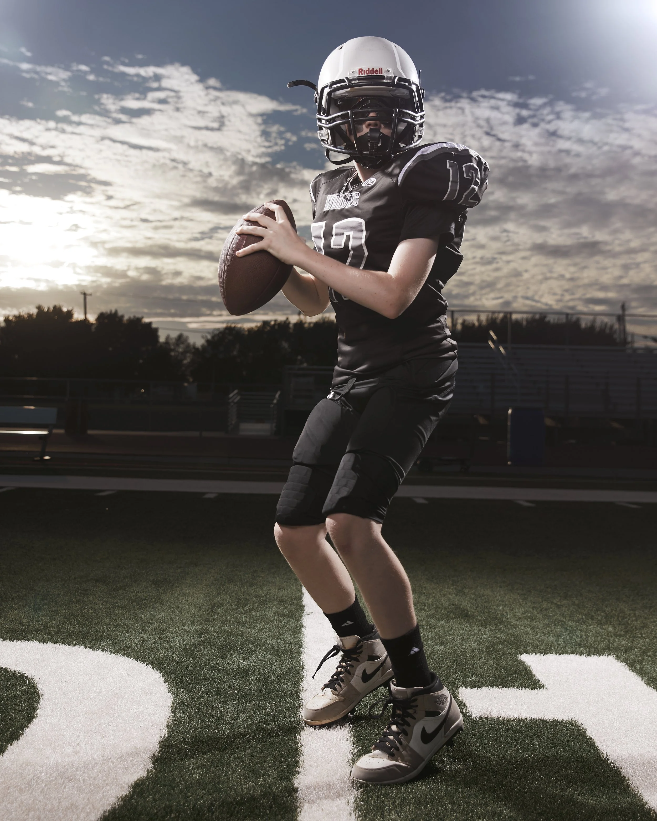 A young football player in black uniform with a helmet holding a football on a football field at sunset or dusk.