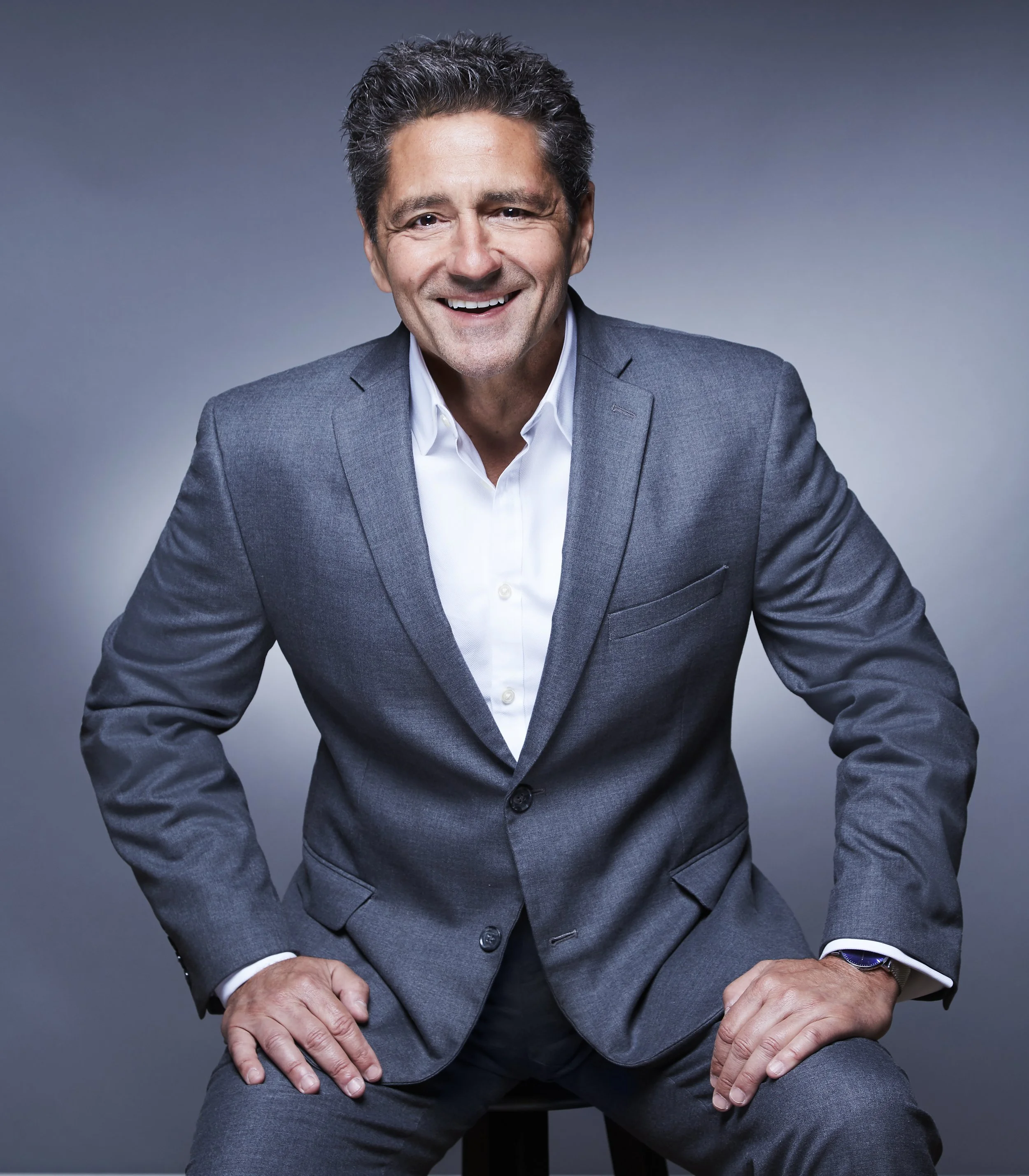 A middle-aged man with dark, curly hair, smiling, wearing a grey suit and white shirt, sitting against a grey background.