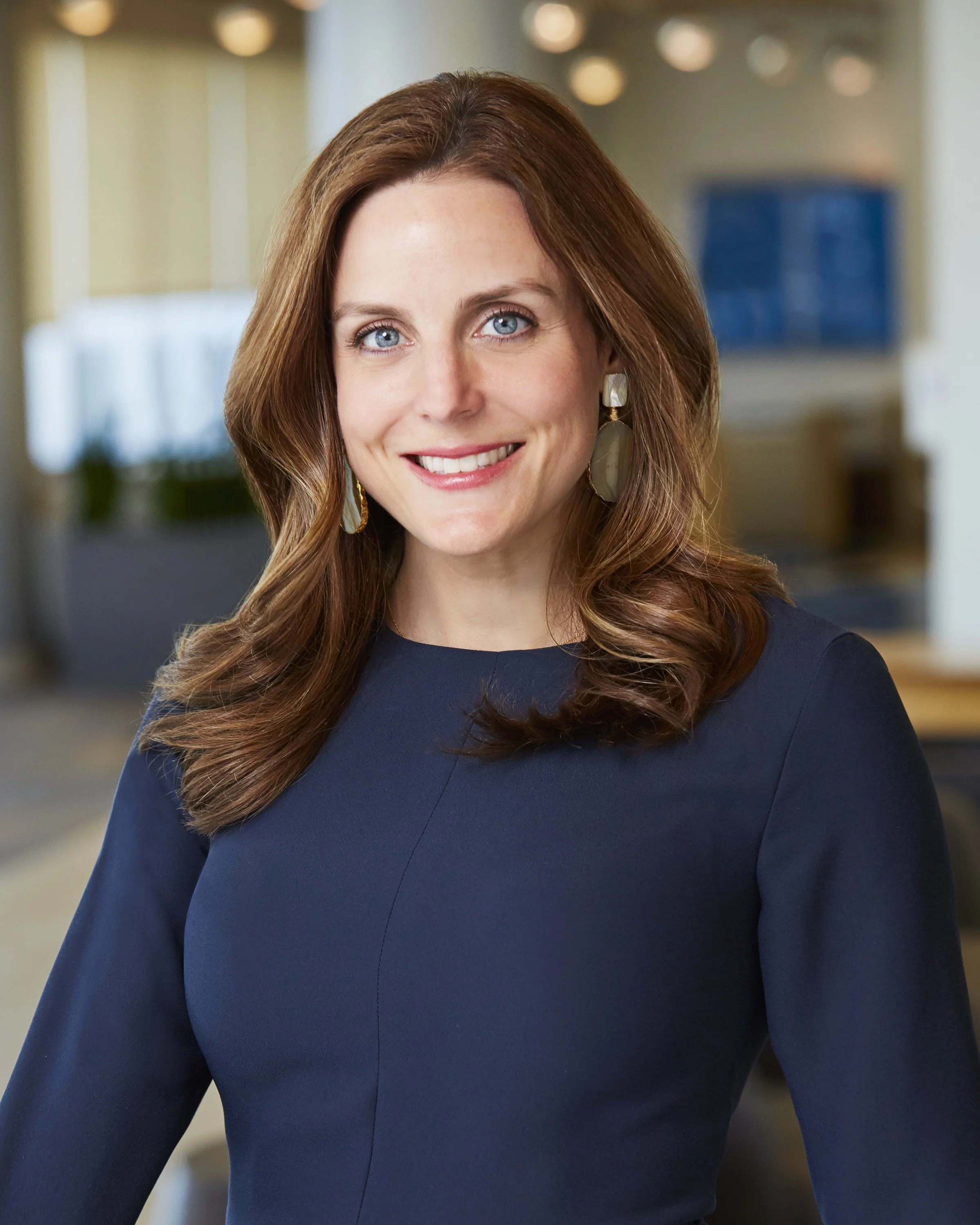 A woman with long red hair, blue eyes, and fair skin, smiling in a dark blue dress with large earrings, in an indoor setting with blurred background.