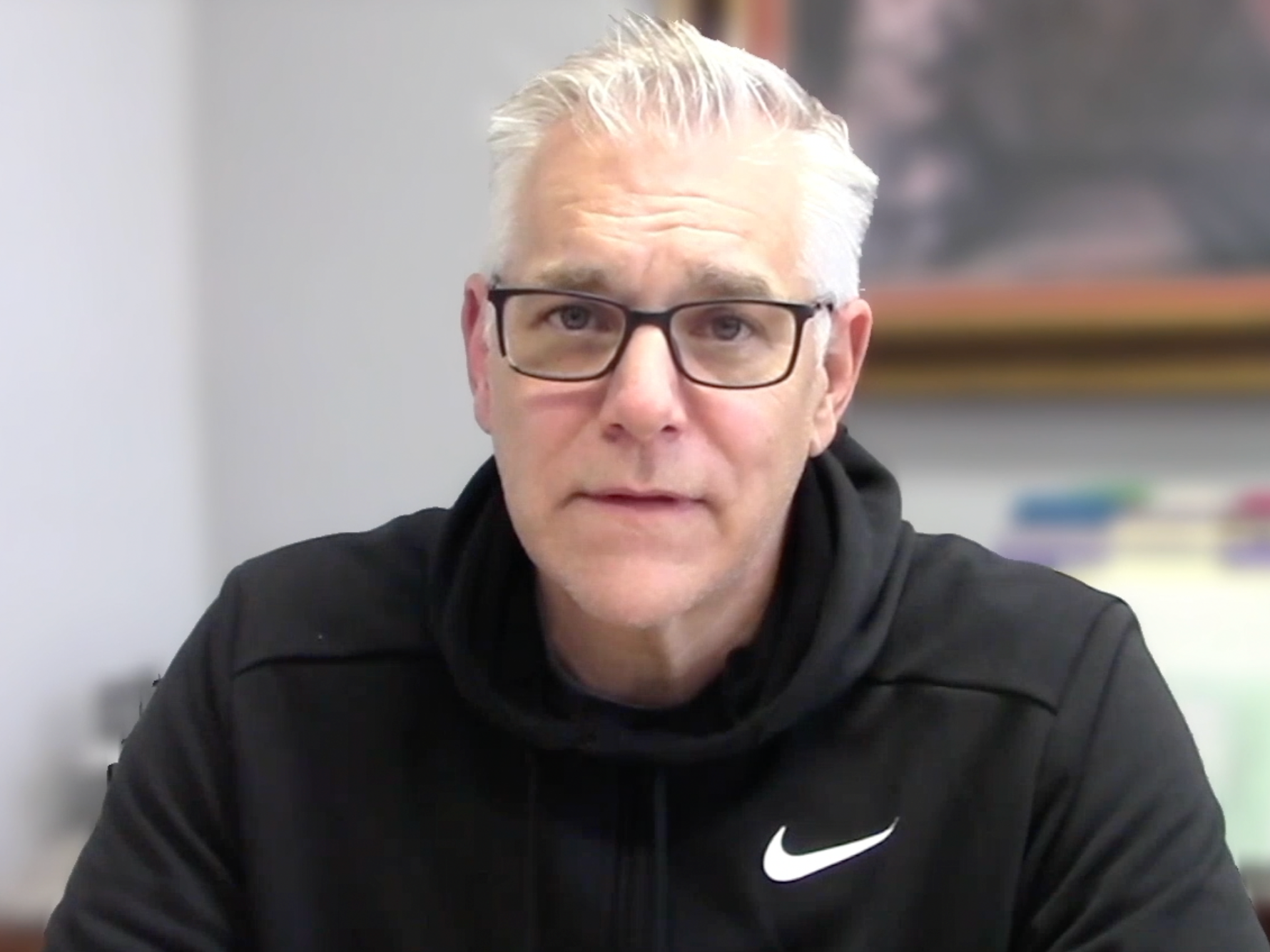 A middle-aged man with gray hair, glasses, and light skin wearing a black Nike hoodie, sitting in an indoor setting with a blurred painting and colorful objects in the background.