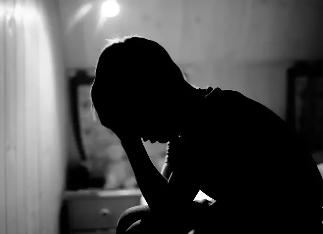 Silhouette of a person sitting with their head bowed and hand on forehead, in a dimly lit room.