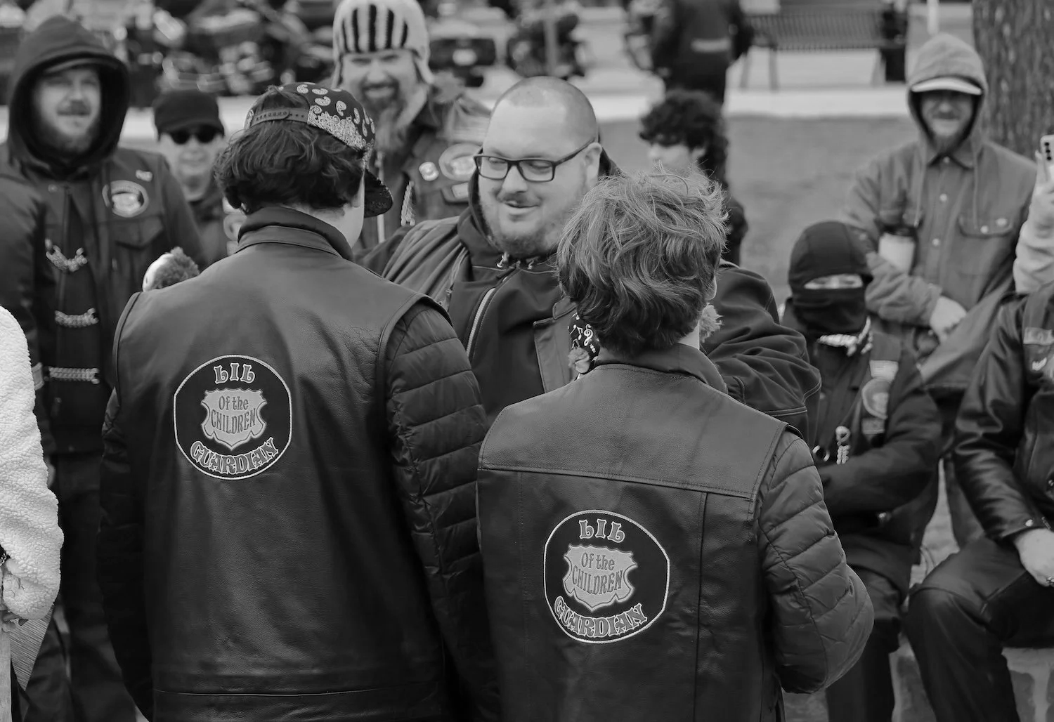 Group of bikers gathered outdoors, some wearing vests with the logo 'Big of the Children', engaging in conversation.