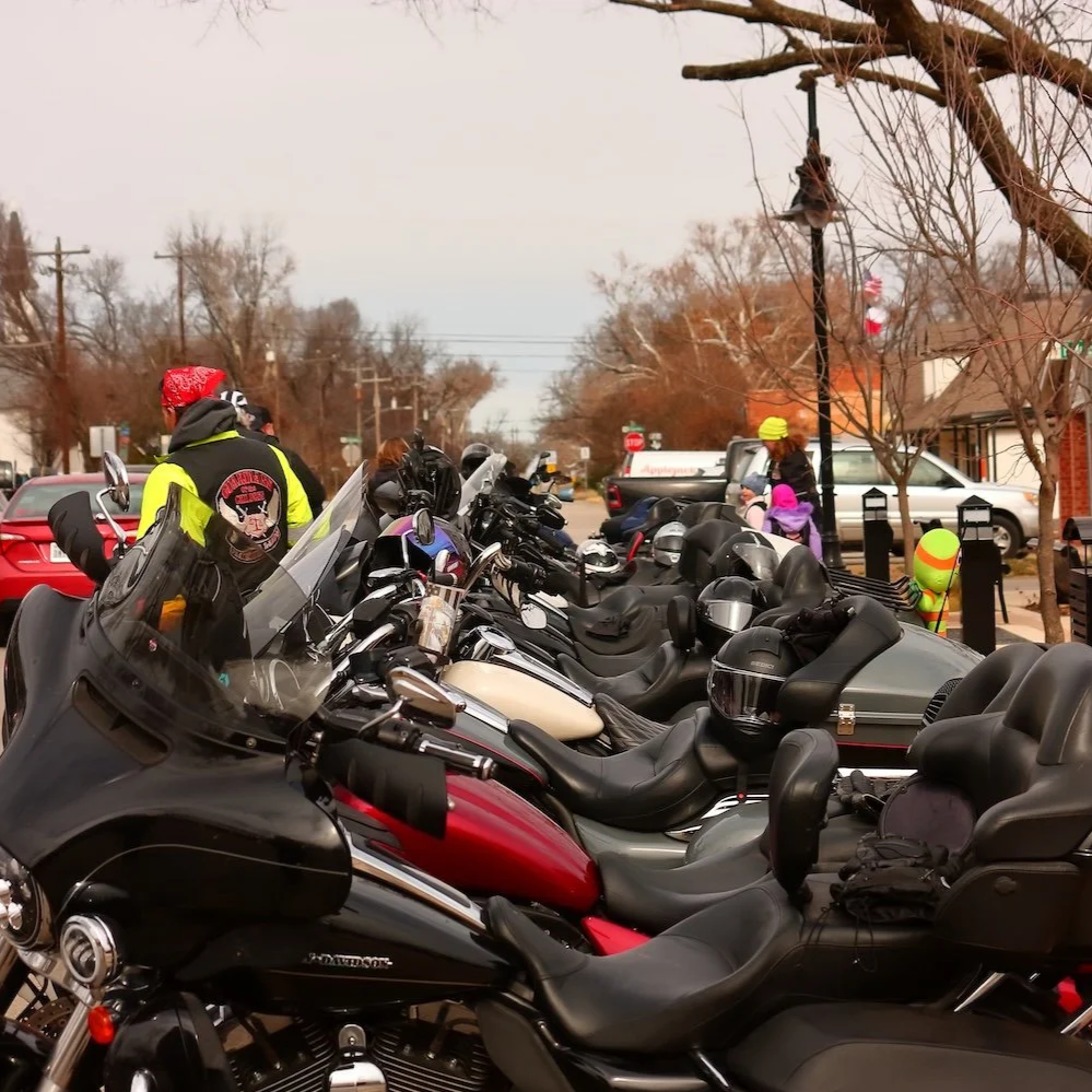 Multiple parked motorcycles in a row on a street. People are seen in the background walking past and talking.