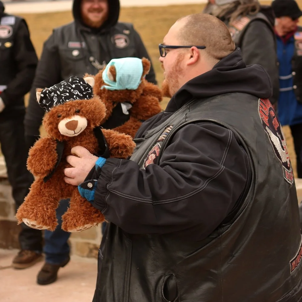 A man in a black leather vest holding a brown teddy bear wearing a bandana and a blue mask, smiling and looking at the teddy bear.