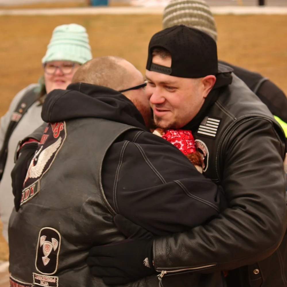 Two men hugging, one with glasses, wearing a black leather jacket and the other with a baseball cap wearing a patch jacket. Two women in the background, one with glasses and a striped hat, the other with a beanie. Indoor setting with wooden backgroun