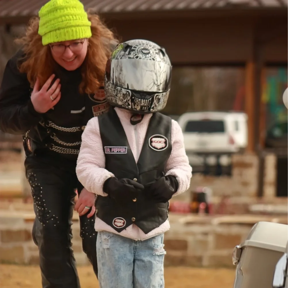 A woman with curly red hair, glasses, and a bright yellow knit hat is smiling and leaning towards a young child. The child is wearing a helmet, a black leather vest with patches, a pink fluffy jacket, gloves, and ripped jeans.