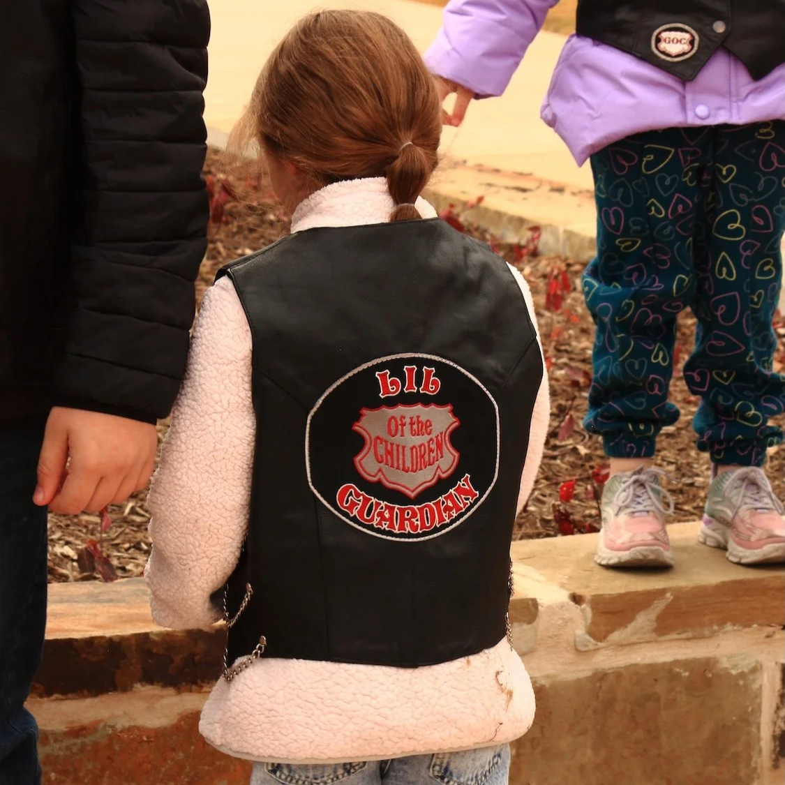 A girl wearing a black vest with an emblem that reads 'LIL GUARDIAN of the CHILDREN' on the back, standing outdoors.