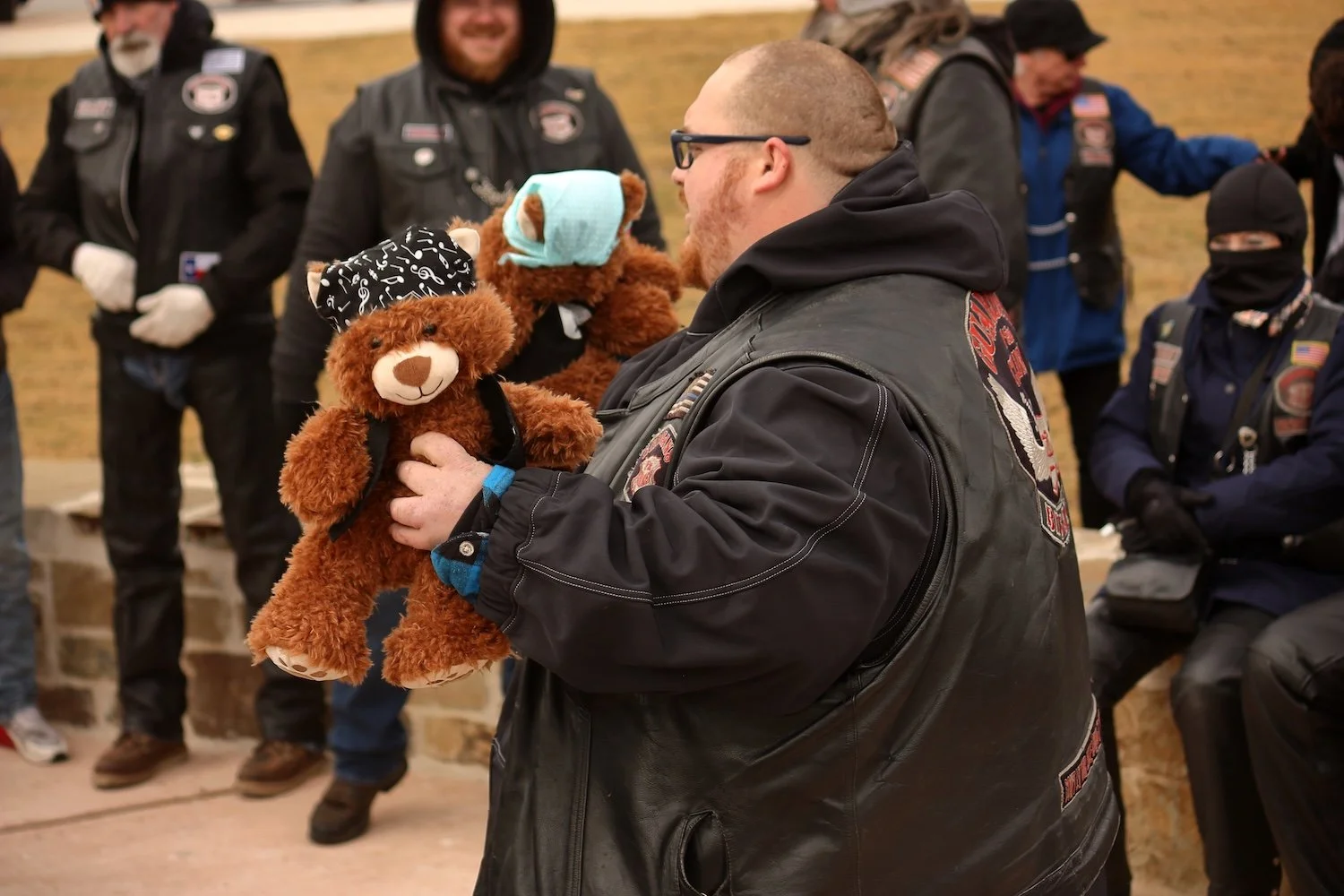 A man in a leather vest holding a teddy bear, surrounded by people dressed in biker gear, Guardian of the Children Big Sandy Chapter.