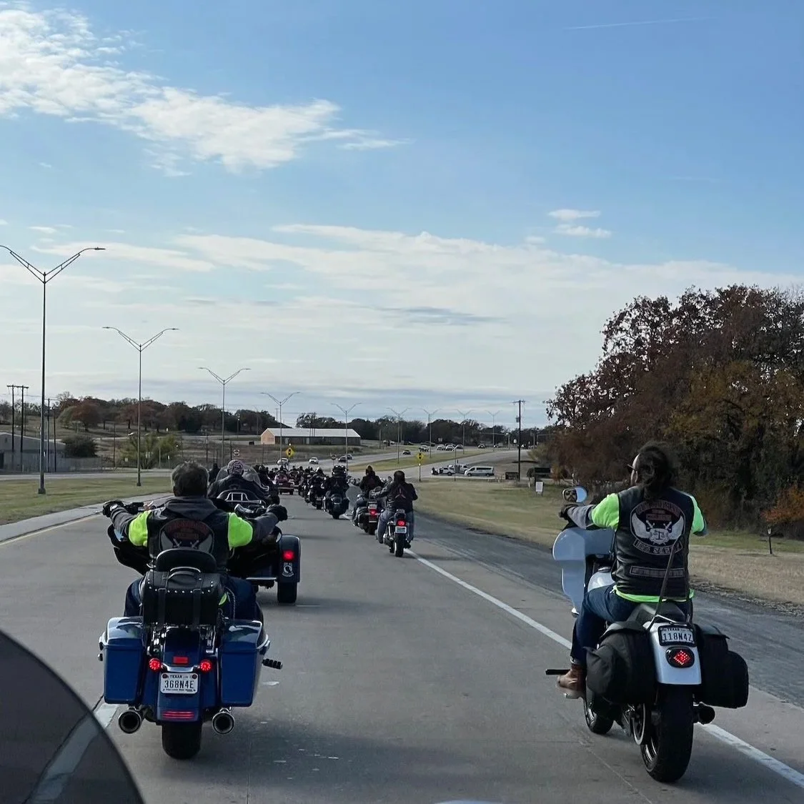 A group of motorcyclists riding on a multi-lane road on a clear day with a blue sky and scattered clouds.