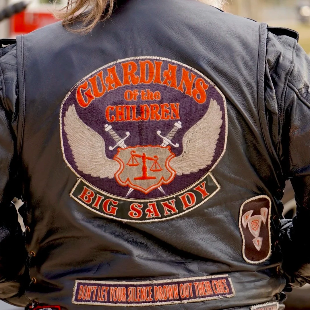 A person wearing a black leather vest with patches, including a large patch that reads "Guardians of the Children" with a winged emblem and swords, and a smaller patch with an R and a shield. There is also a slogan patch at the bottom that says, "Don't let your silence drown out their cries."