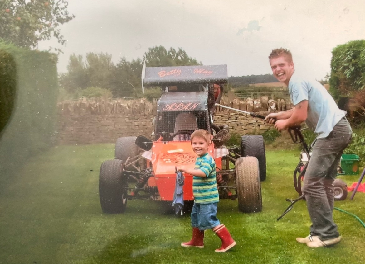 A man and a young boy washing a race car together in a backyard on a cloudy day.