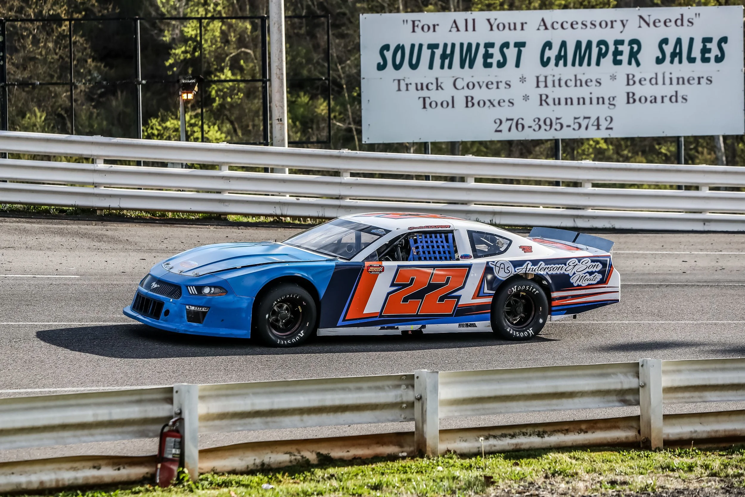 A race car with the number 22 painted on its side driving on a race track. The car is mainly blue and white with orange accents. There is a billboard in the background advertising campground supplies, including truck covers, hitches, bedliners, toolb