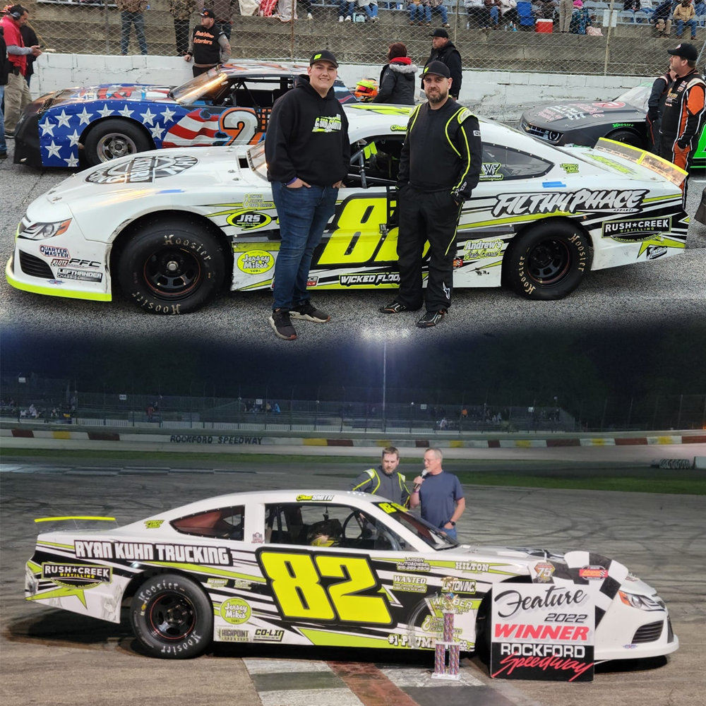 Two stock cars at a race track; one car held by two men, indicating a win, with trophies and a congratulatory sign, and the other car with two men standing beside it, with a crowd in the background.