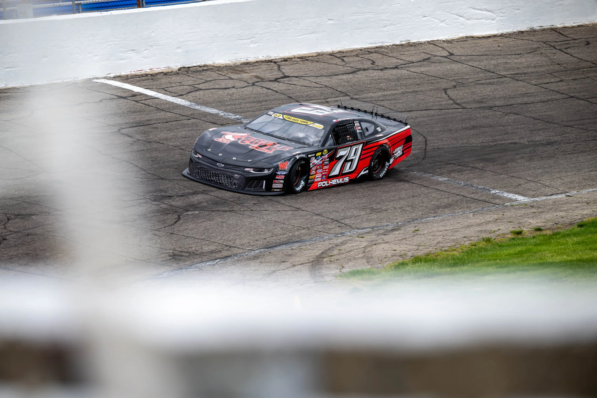 Black and red race car with the number 79 on the side, on a racetrack with paint and cracks, surrounded by white barriers.
