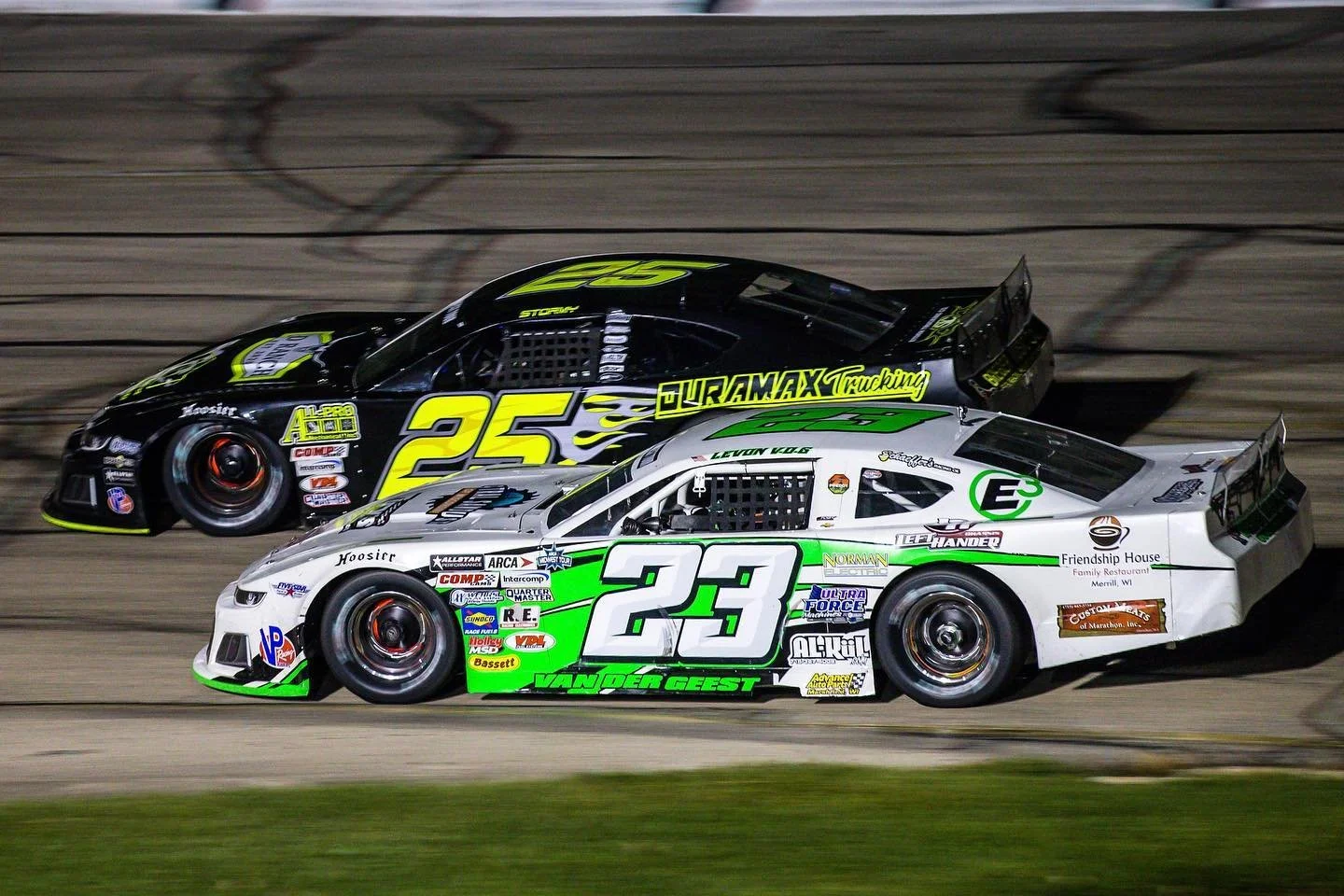 Two race cars competing on a dirt track at night. One car is black with yellow accents and the number 25, the other is white with green accents and the number 23.
