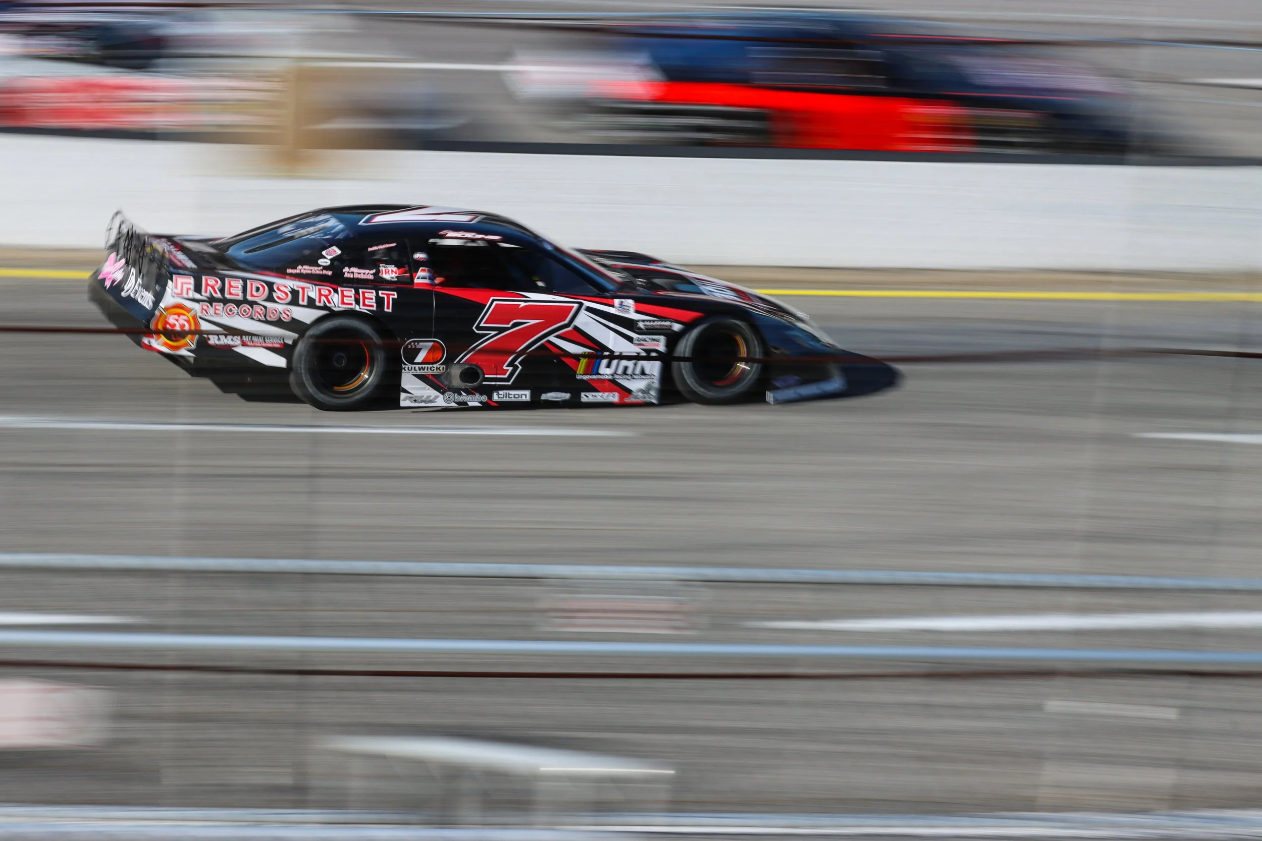 Black racing car with the number 7 and red and white decals speeding on a race track, with a blurred background indicating high speed.
