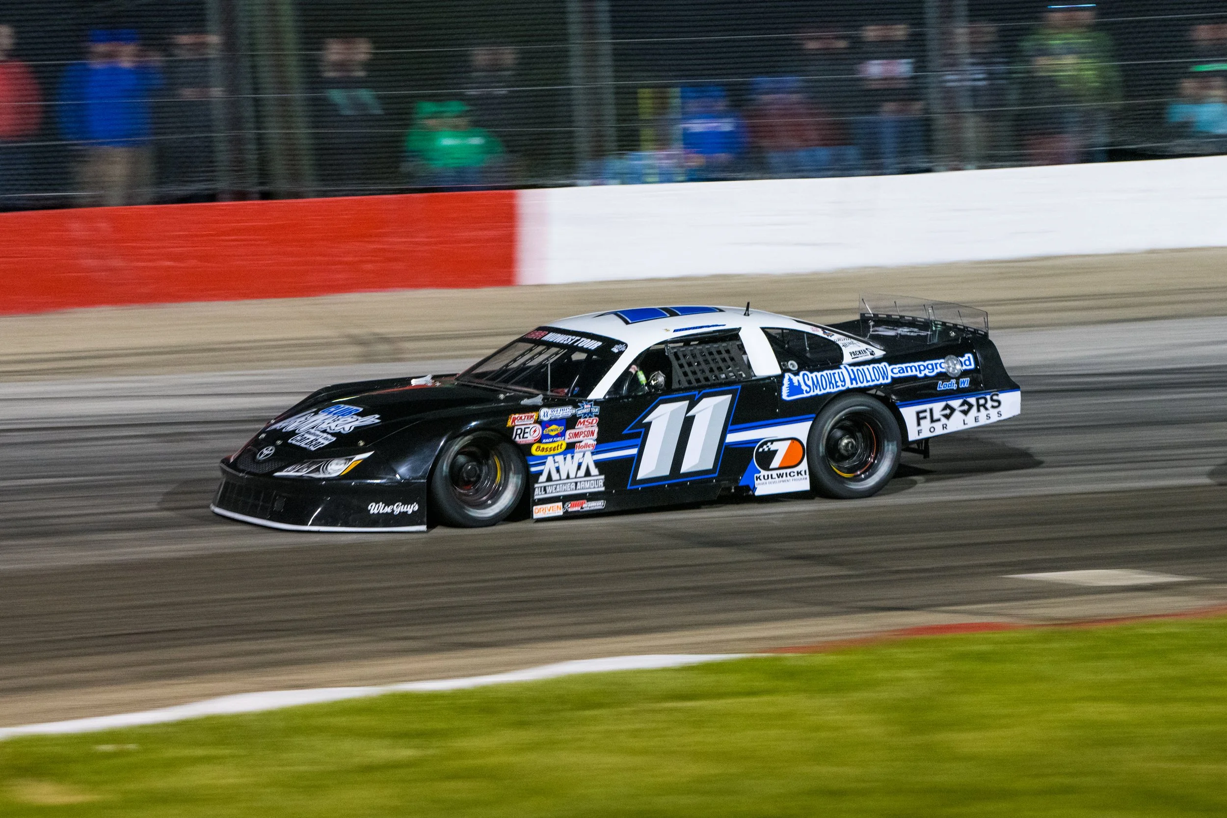A black and white race car with the number 11 on its side speeds along a dirt track at night, with spectators watching behind a fence.