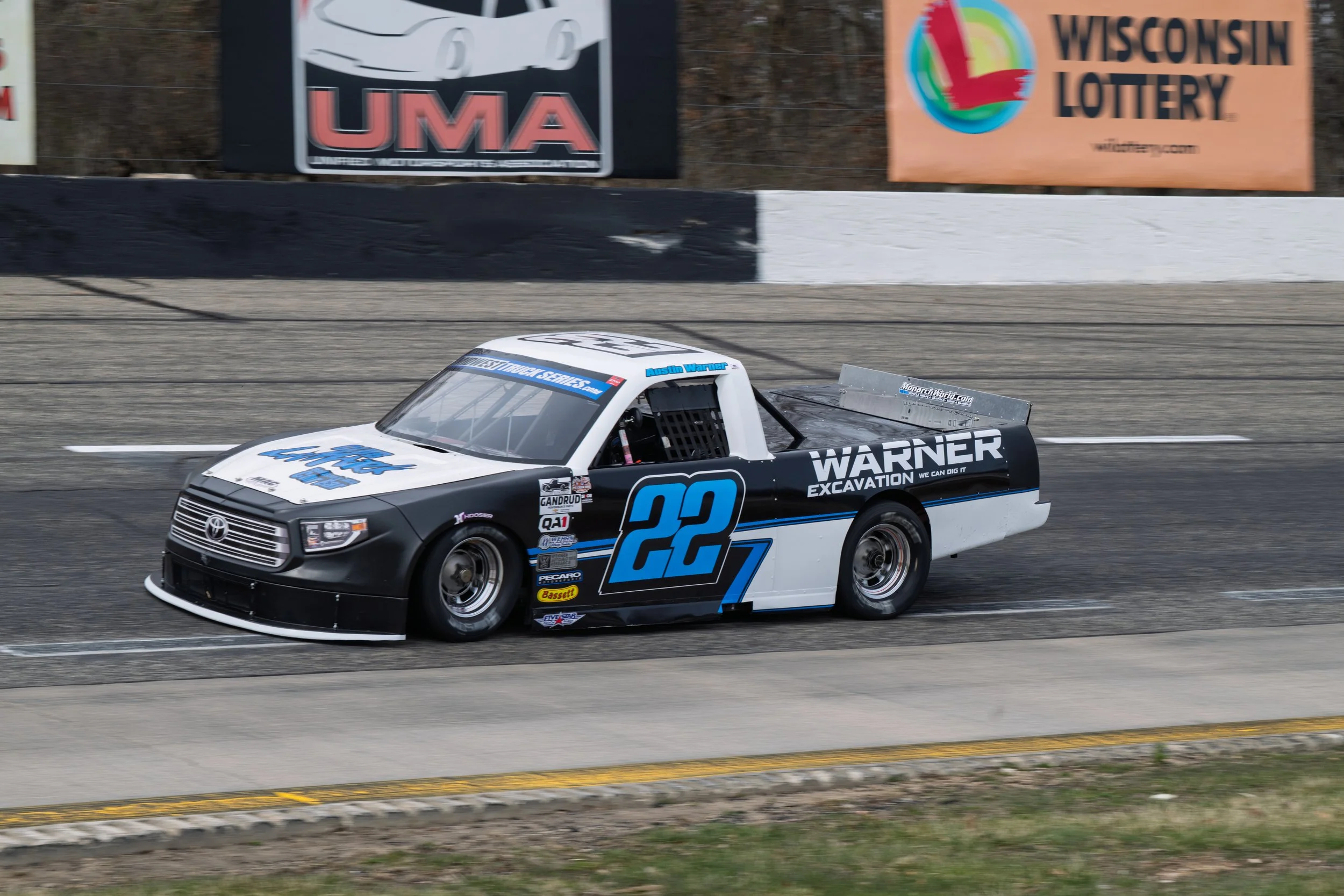 A black and white race car with blue accents, number 22, speeding on a race track. The car has sponsor logos including Warner Excavation. Background includes advertising signs for Wisconsin Lottery and UMA.