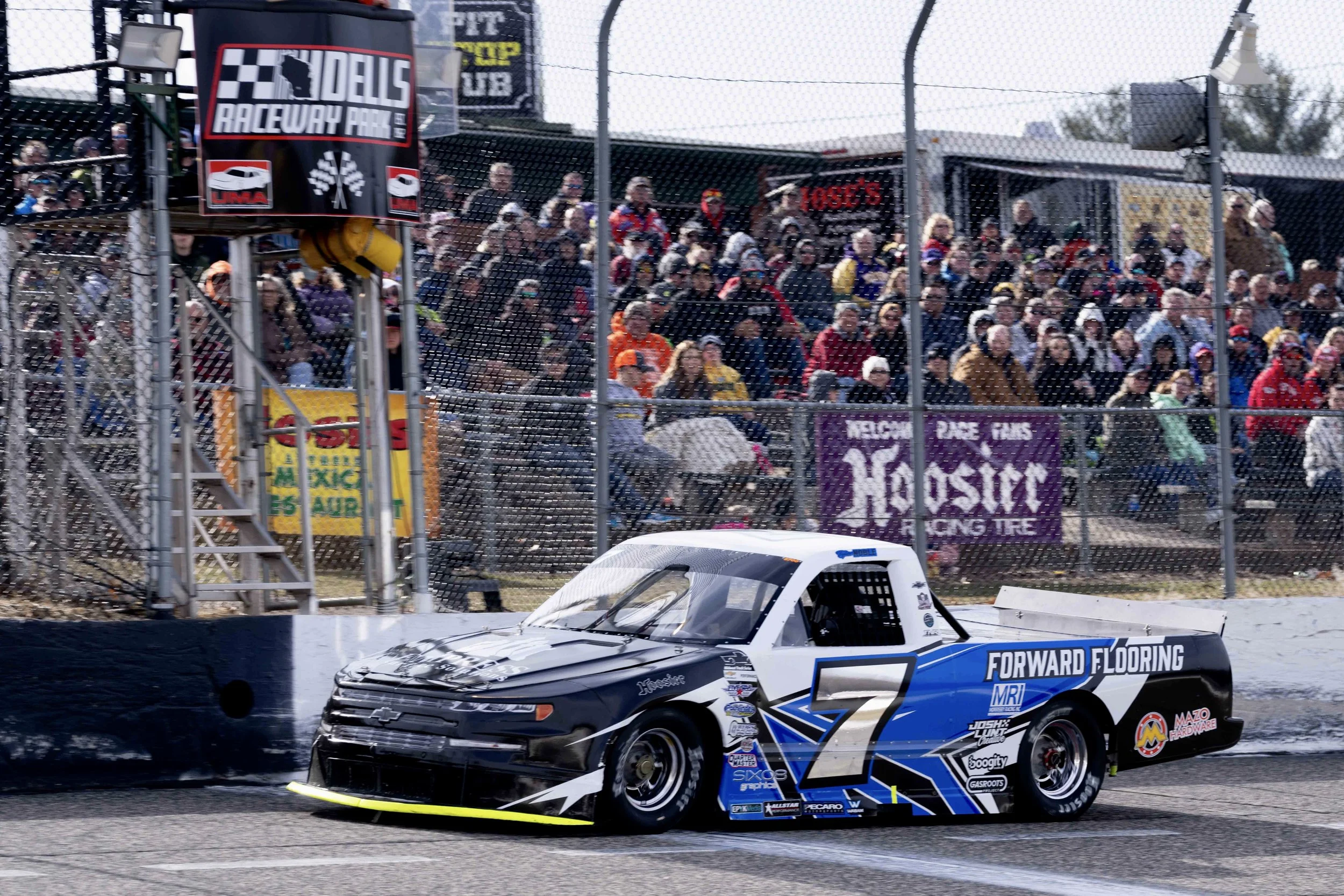A race car numbered 7 driven by Chevrolet, speeding on a racetrack near a crowd of spectators behind a fence.