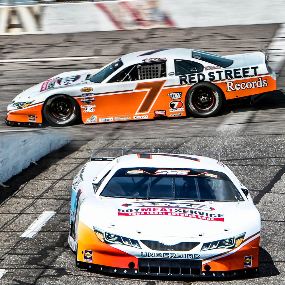 Two race cars on a racetrack, one in front of the other, with motion blur in the background. The cars are white with orange and black accents and sponsorship logos.