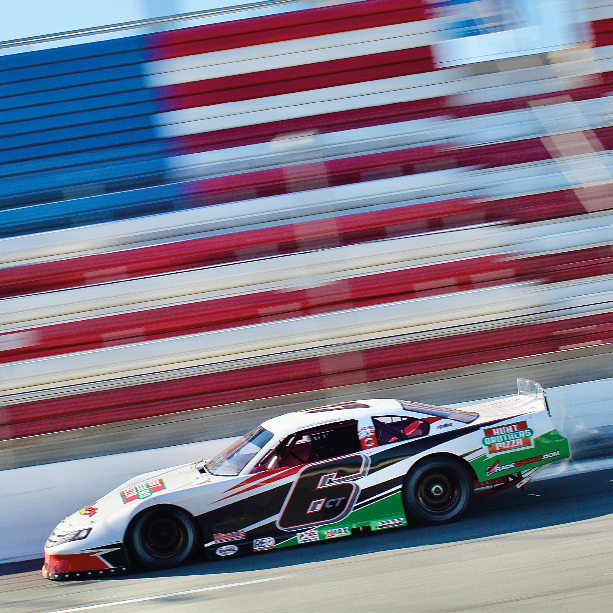 A race car with a black, white, red, and green design speeds on a racetrack, with red, white, and blue bleacher seats blurred in the background.