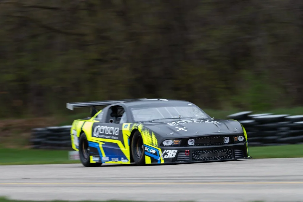A black and yellow race car speeding on a track with a blurred background and tire barriers on the side.