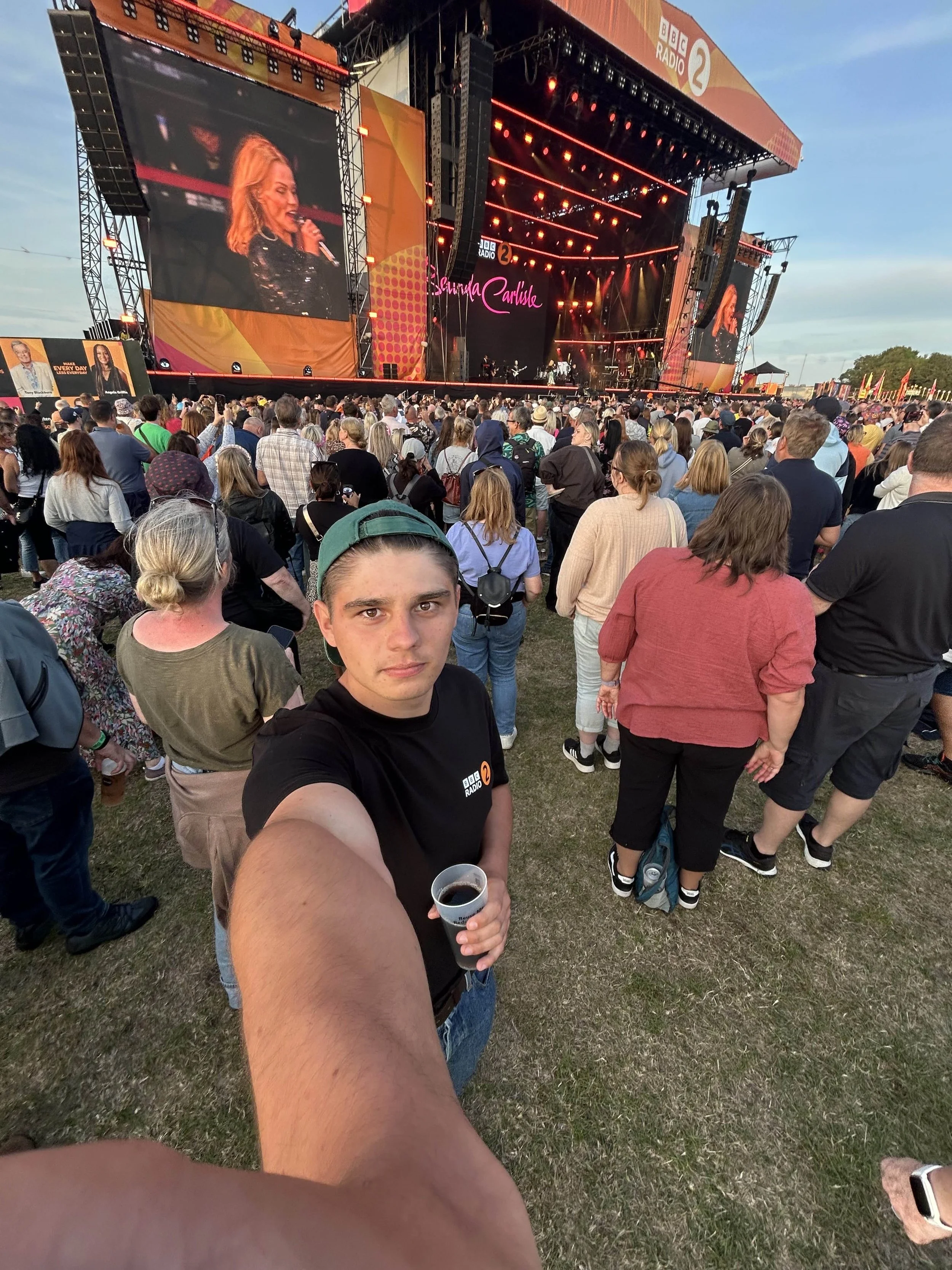 A crowd of people attending an outdoor concert or festival in the evening. The stage has large screens displaying a female singer with blonde hair, wearing a dark outfit. The stage is adorned with bright lights and branding including BBC Radio 2. The person in the foreground is taking a selfie, holding a drink, and wearing a black BBC Radio 2 T-shirt.