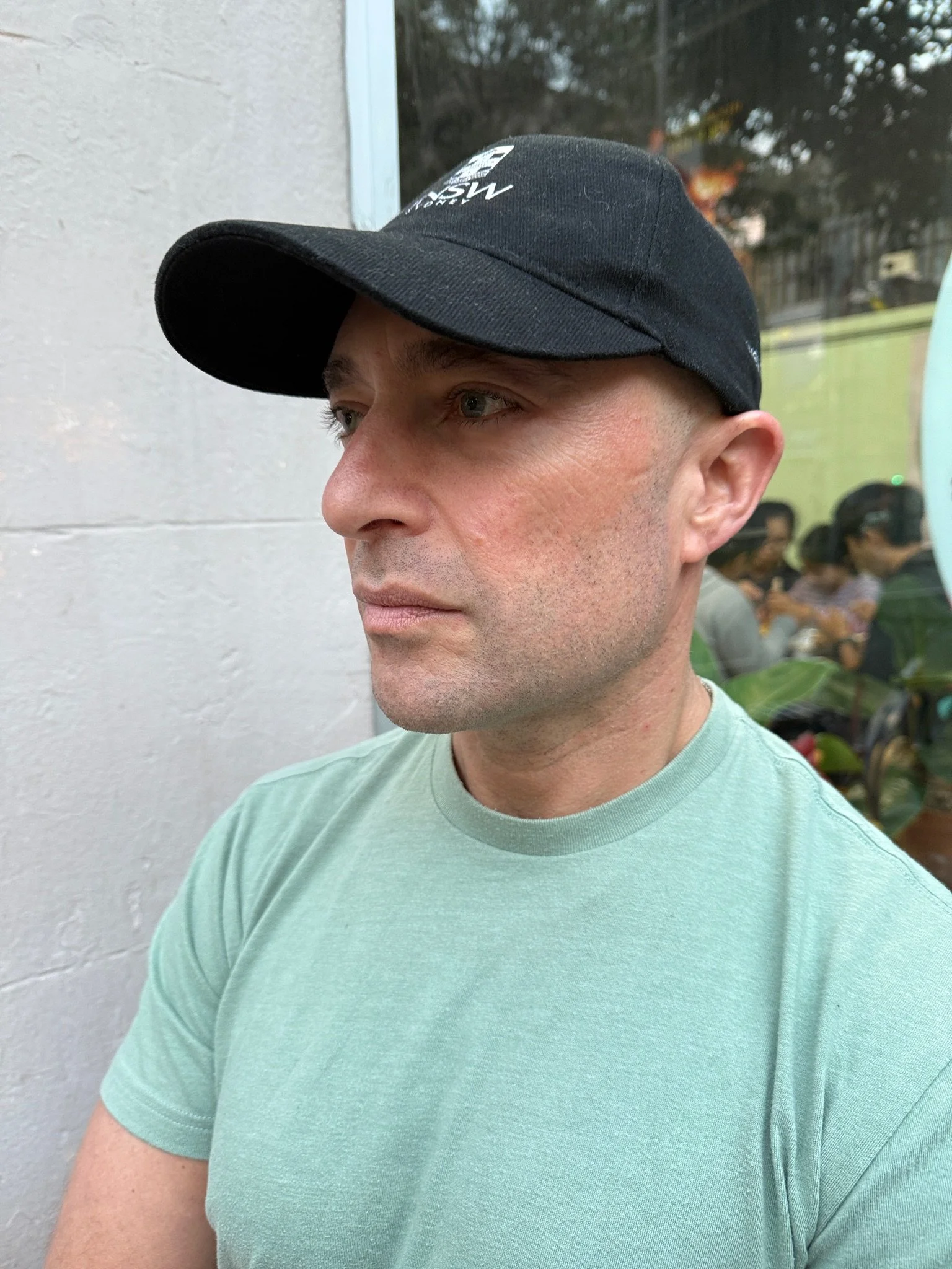 A man wearing a black cap and a light green t-shirt, standing against a white wall with a mirror and some people in the background.