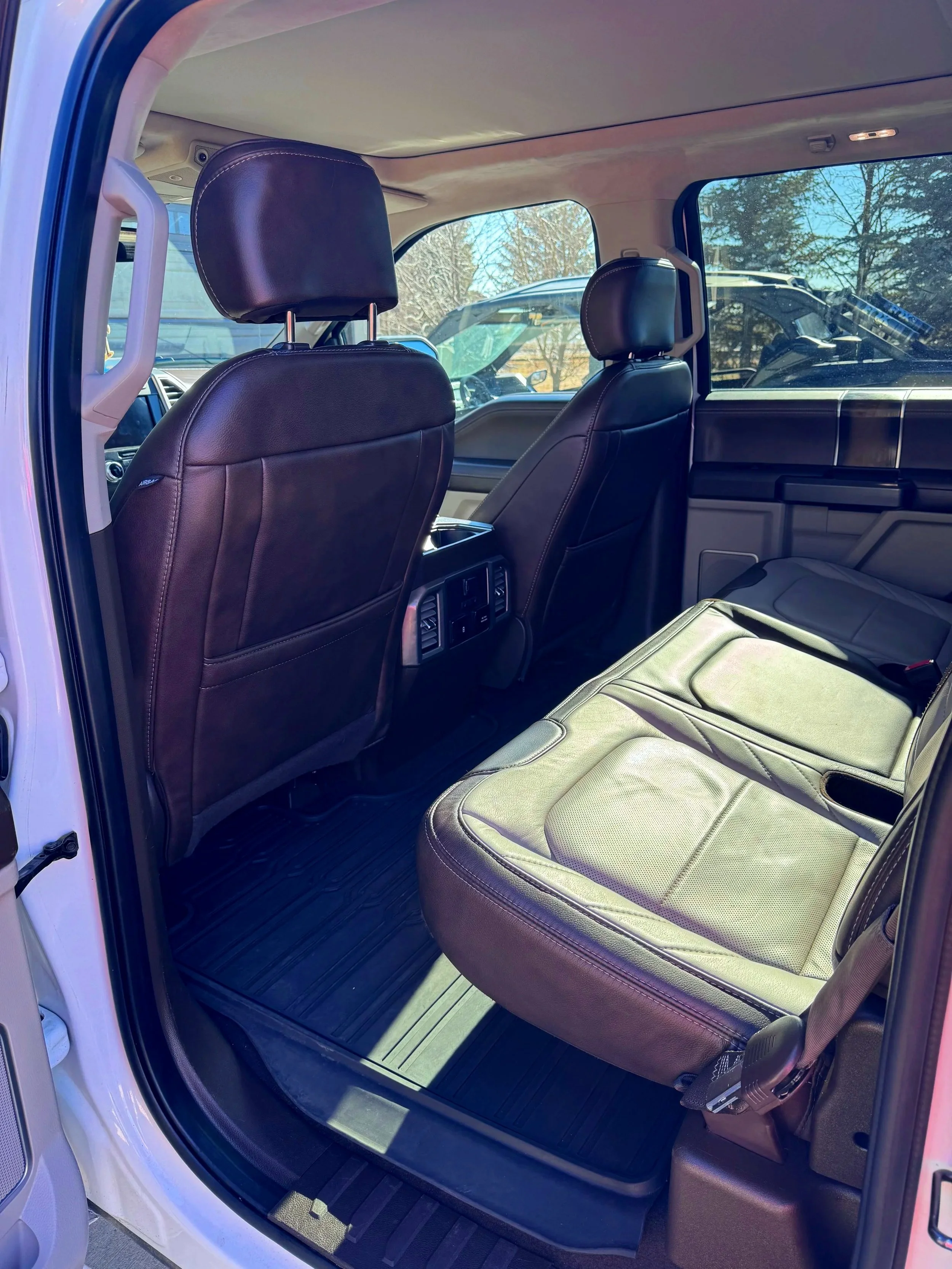 The interior rear passenger area of a white truck with black leather seats and floor mats. Outside the vehicle, trees and other vehicles are visible through the windows.