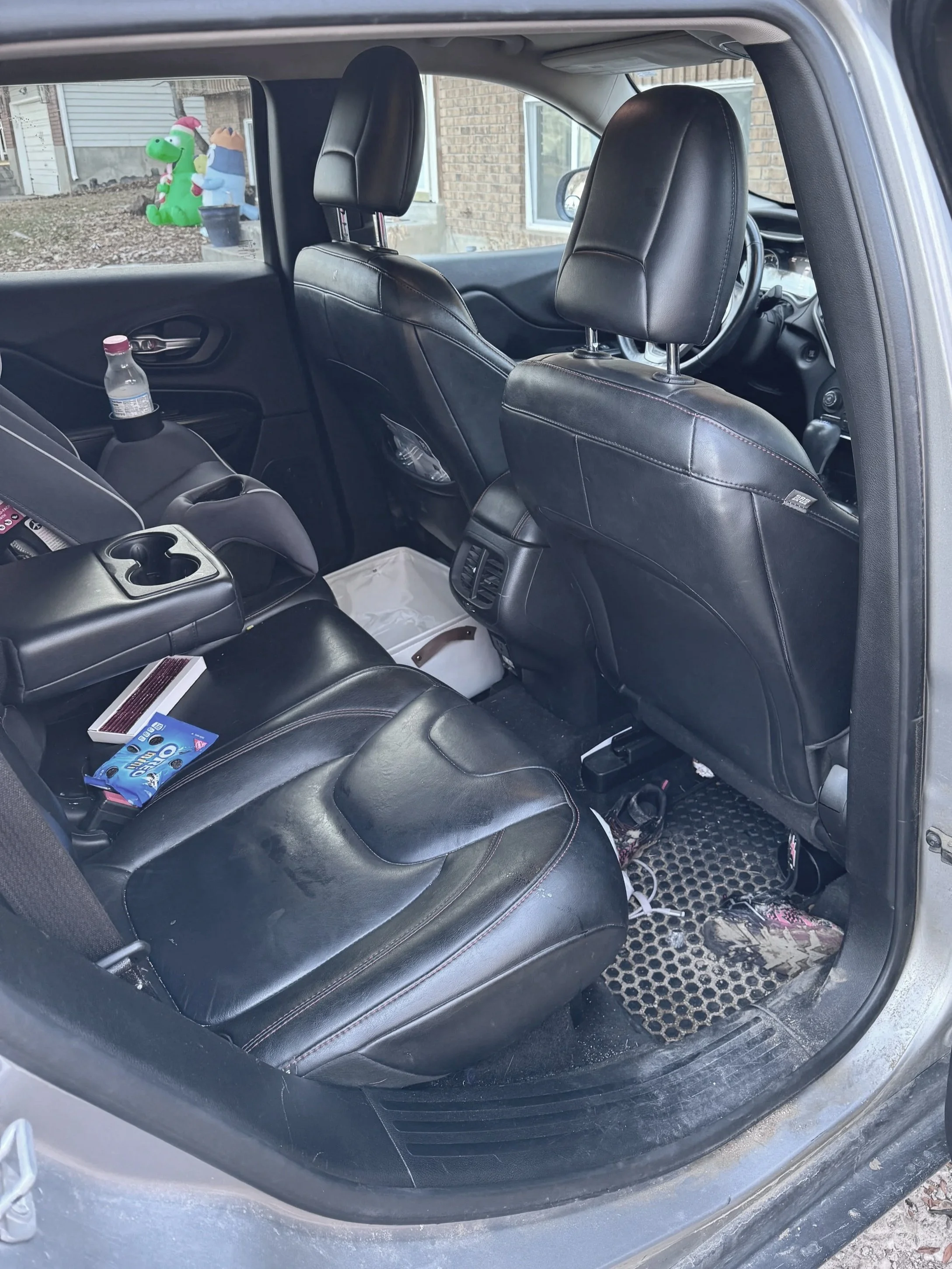 View of the back seat area inside a car, showing black leather seats, a water bottle in the side pocket, and various items on the seat and floor, with a window showing a yard with inflatable decorations outside.