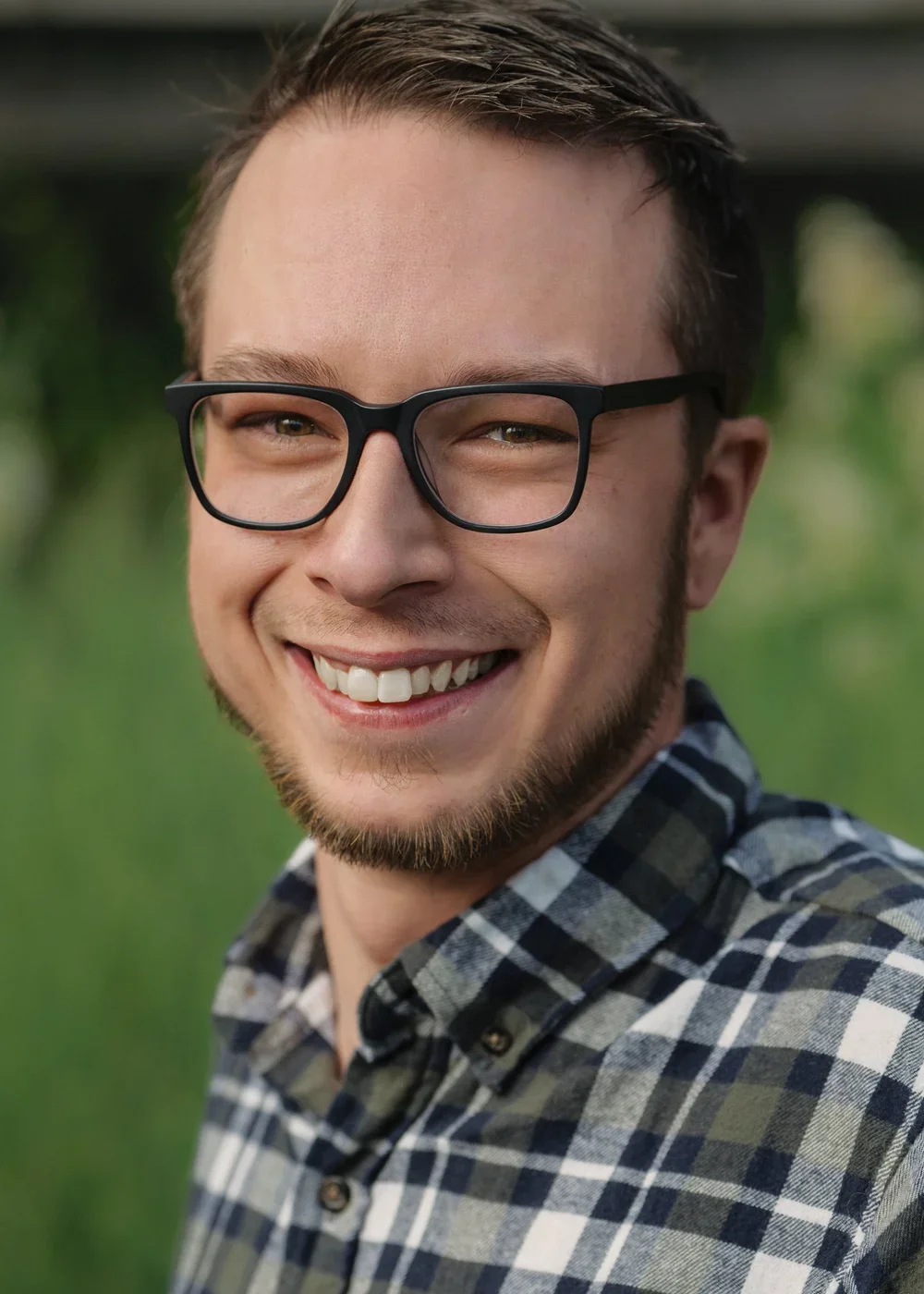 A young man smiling outdoors, wearing black-rimmed glasses and a plaid shirt, with a blurred green background.