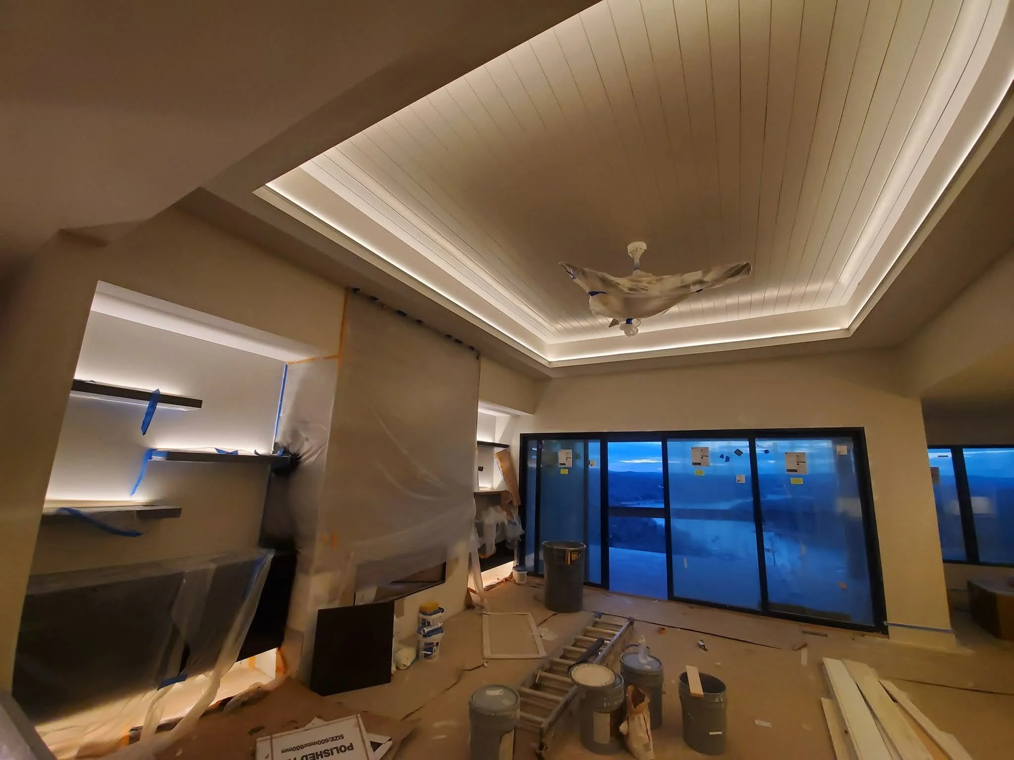 Interior of a building under construction or renovation, with a ceiling equipped with recessed mood lighting, and constructing materials and tools scattered around the floor, including buckets, wood pieces, and construction boxes.