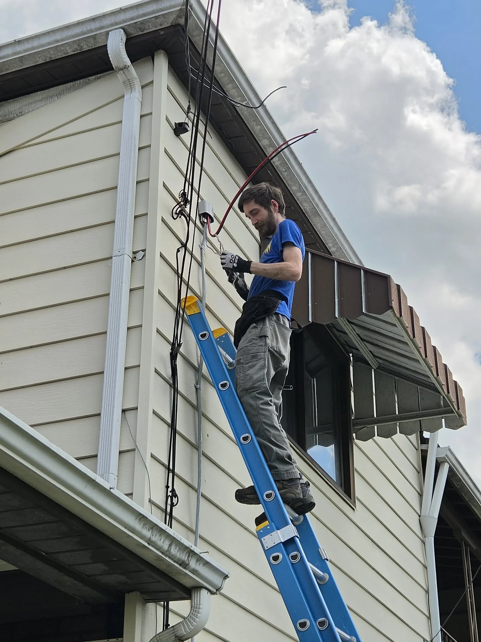 A man with a beard, wearing gloves, a blue shirt, and gray pants, is standing on a blue ladder on the side of a house working on electrical wiring or a cable connection on the house's exterior. The house has beige siding and a gutter system, and the sky is partly cloudy.