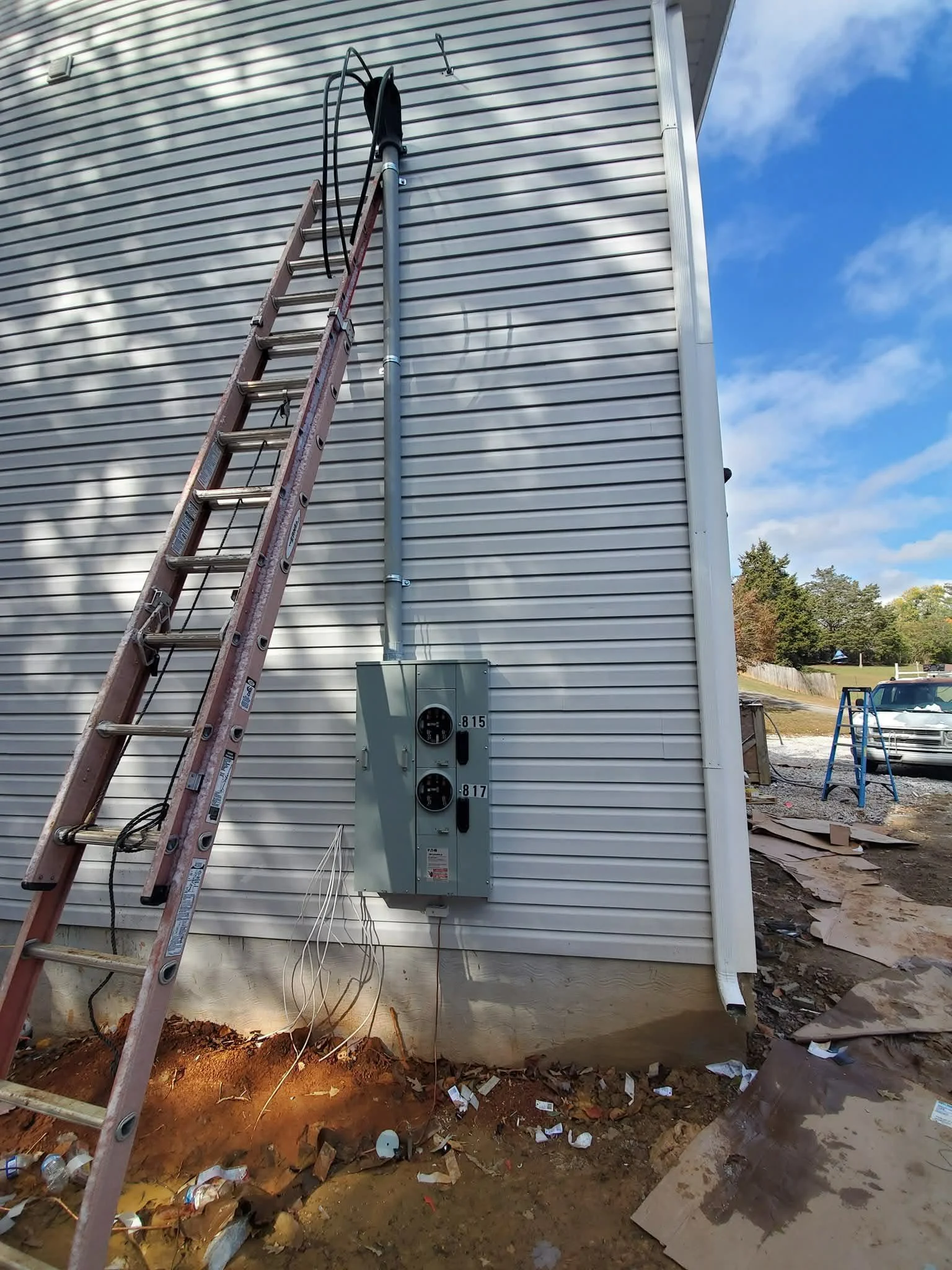 Exterior wall of a building with a ladder leaning against it, electrical meters, and conduit pipes. Construction debris and dirt on the ground, with some cars and trees in the background and a partly cloudy sky.