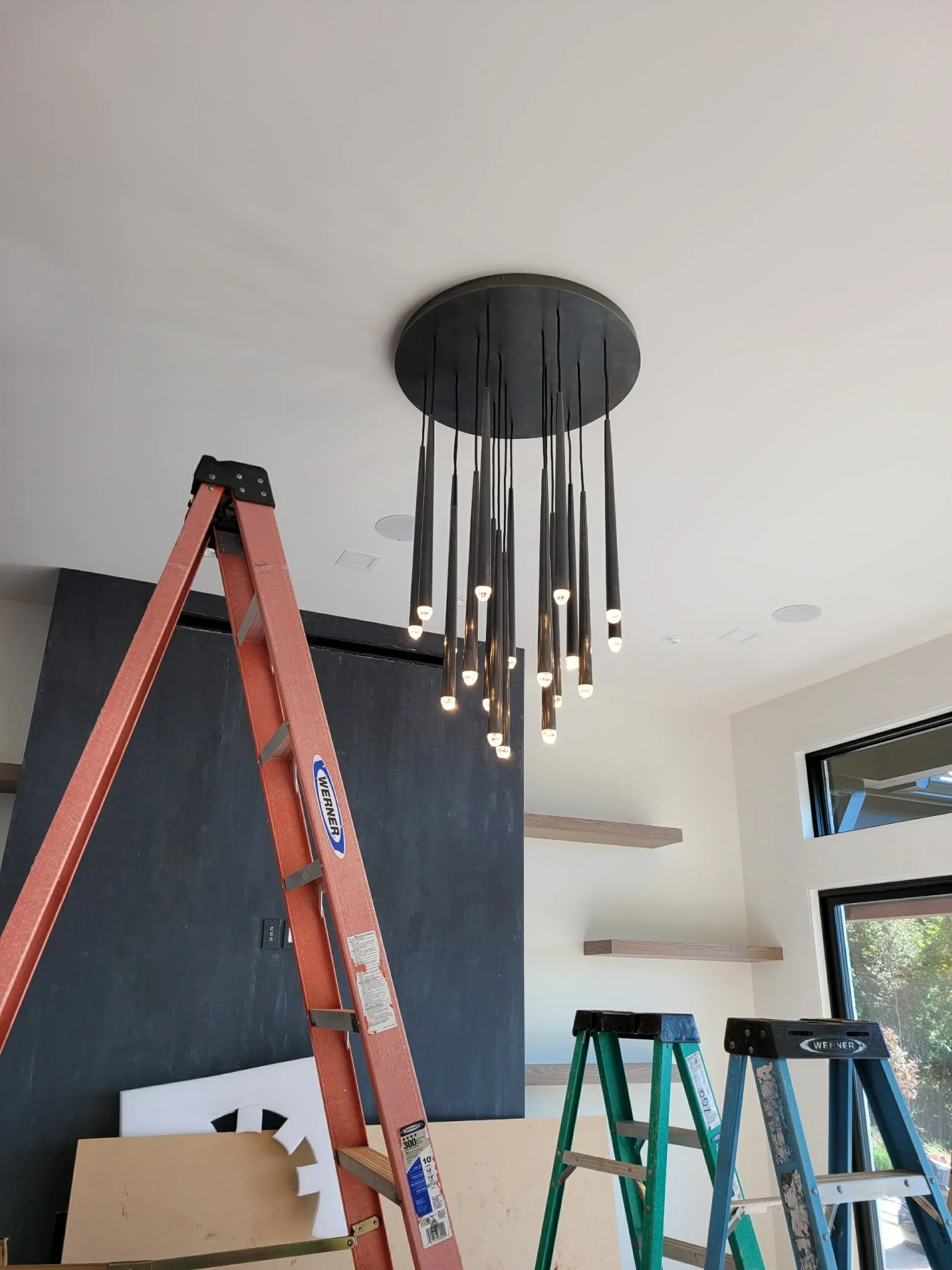 Interior view of a room under renovation, featuring a modern chandelier hanging from the ceiling, several ladders in different colors and sizes, a black accent wall, and windows letting in natural light.
