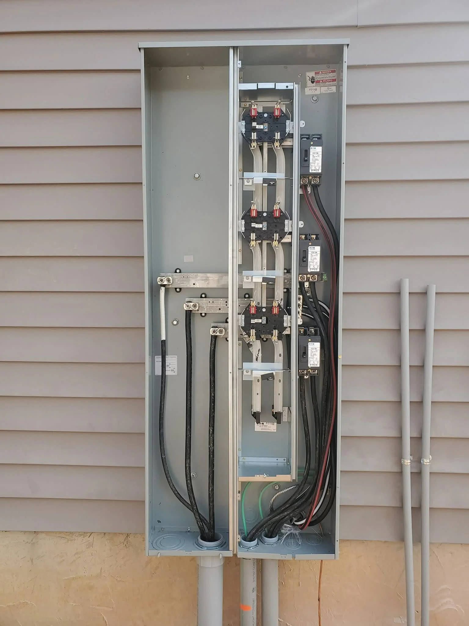 An empty electrical meter box with wiring and circuit breakers, mounted on a beige wall outside a house.