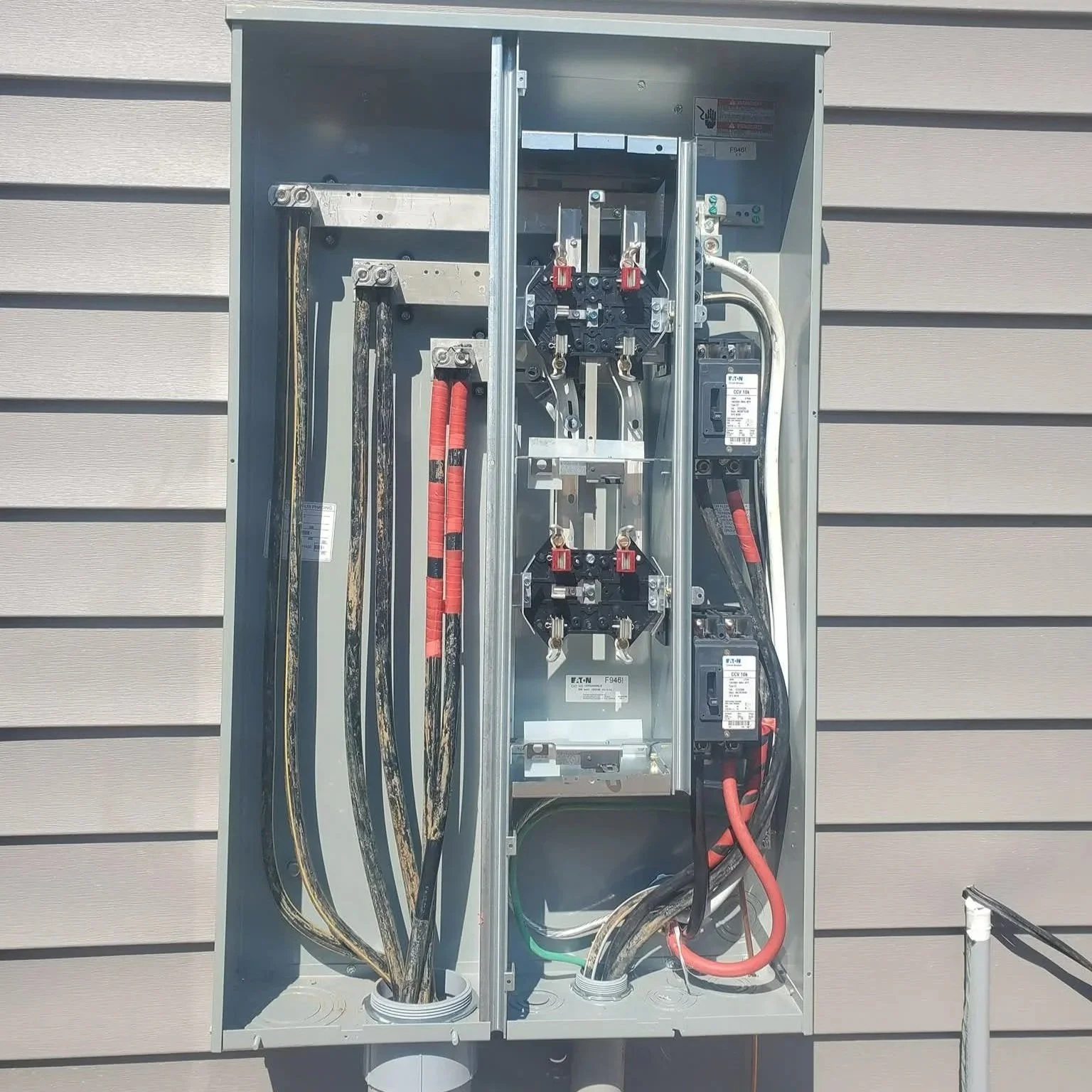 Open electrical utility box with various electrical cables and components mounted inside, located outside on a beige siding wall.