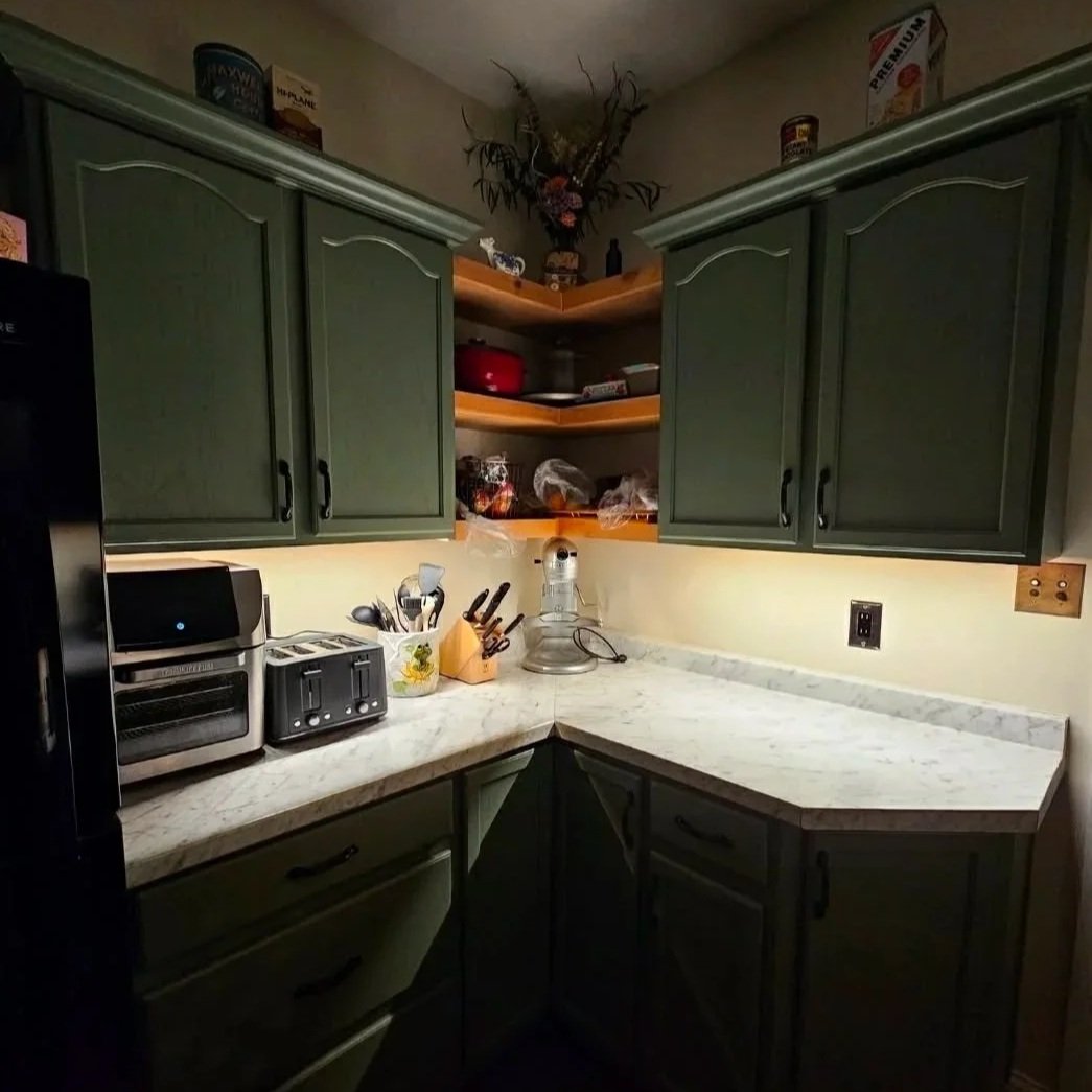 A small kitchen corner with green cabinets, a marble countertop, and open wooden shelves holding various kitchen items. Counters are lit by under cabinet lighting.