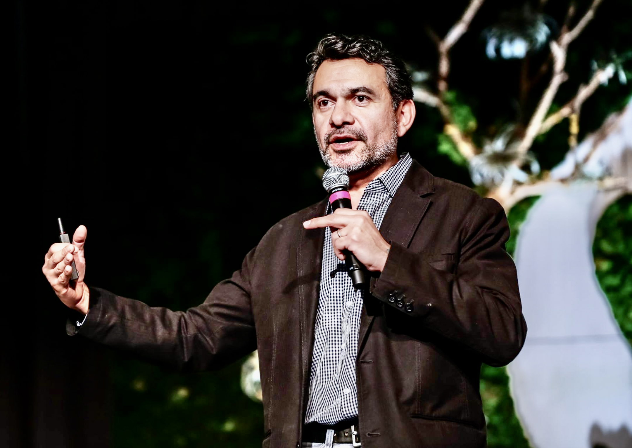 A man with dark hair and a beard giving a presentation indoors. He is holding a microphone in his right hand and a clicker in his left hand, wearing a brown blazer and a checkered shirt.